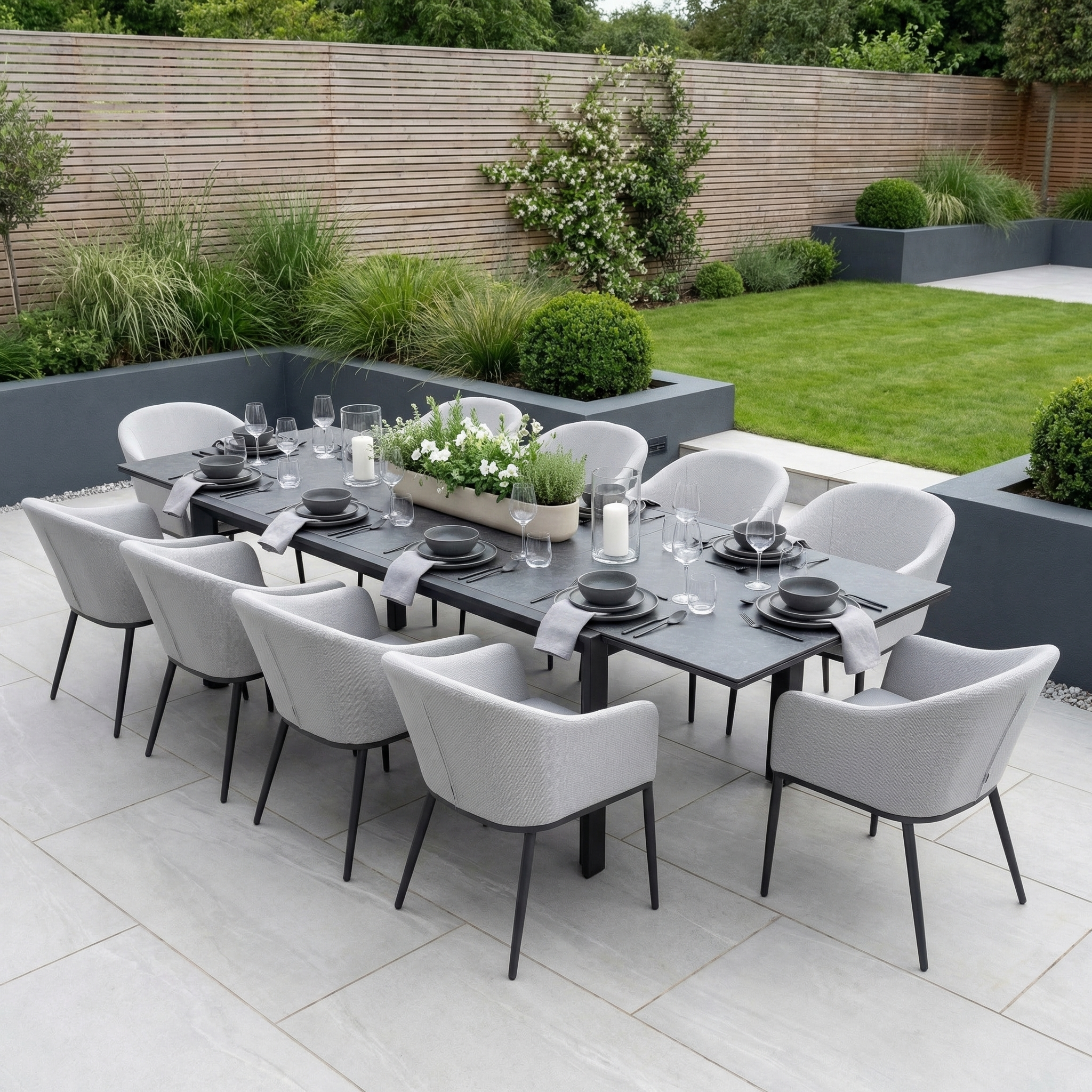 The Luna 10 Seat Outdoor Fabric Extending Ceramic Dining Set in Oyster Grey is perfect for patios, offering modern all-weather comfort surrounded by lush garden landscaping.
