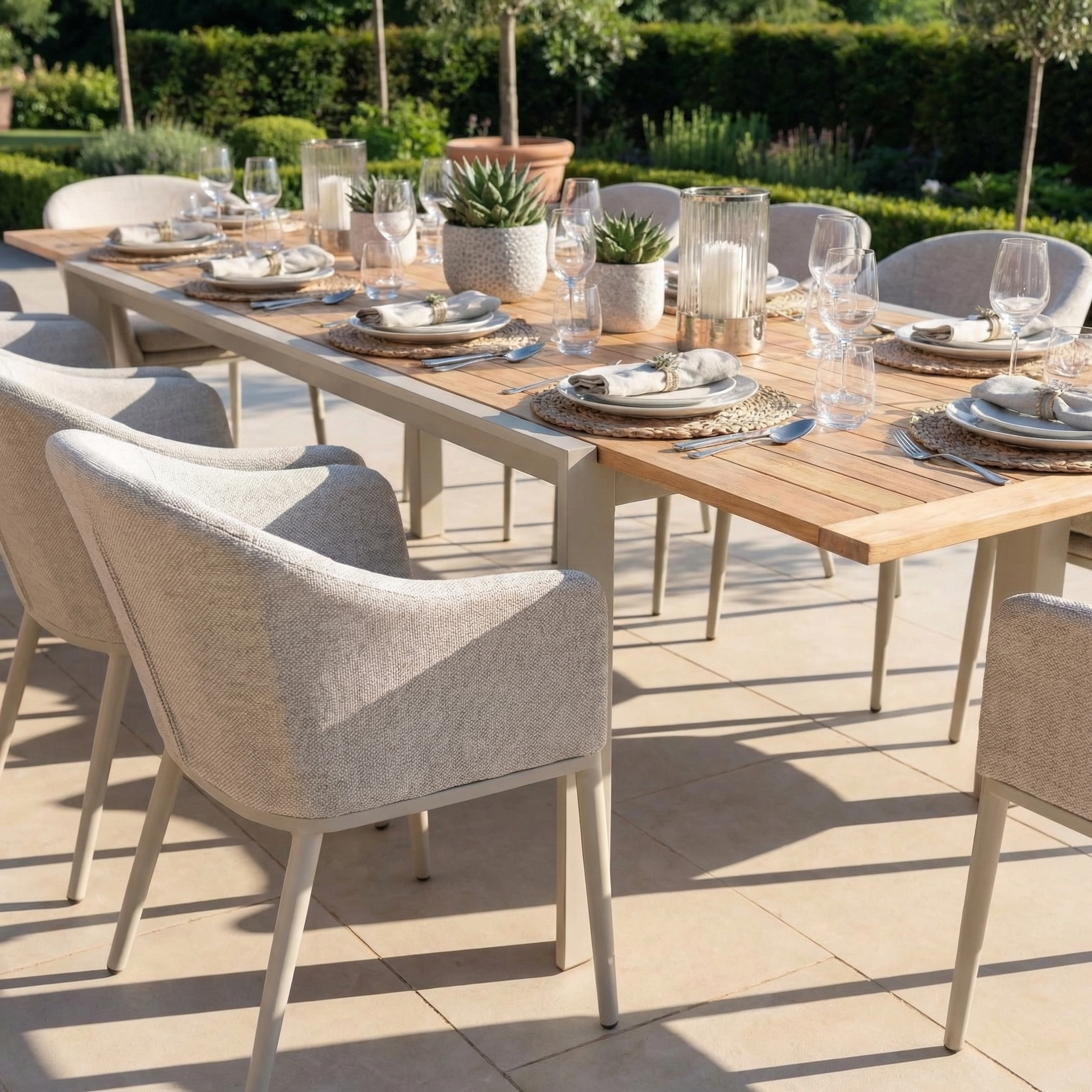 The Luna 10 Seat Outdoor Fabric Extending Teak Dining Set in Fawn is arranged on a sunny patio, topped with plates, glasses, candles, and potted plants for a stylish outdoor dining experience.