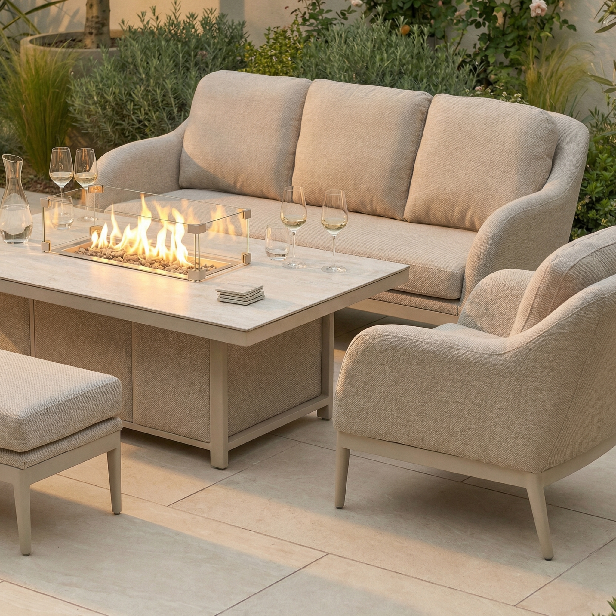 The Luna 3 Seat Outdoor Fabric Sofa Set with Rising Firepit Table in Fawn is crafted from durable all-weather material and arranged for outdoor gatherings, shown with wine glasses and plants in the background.