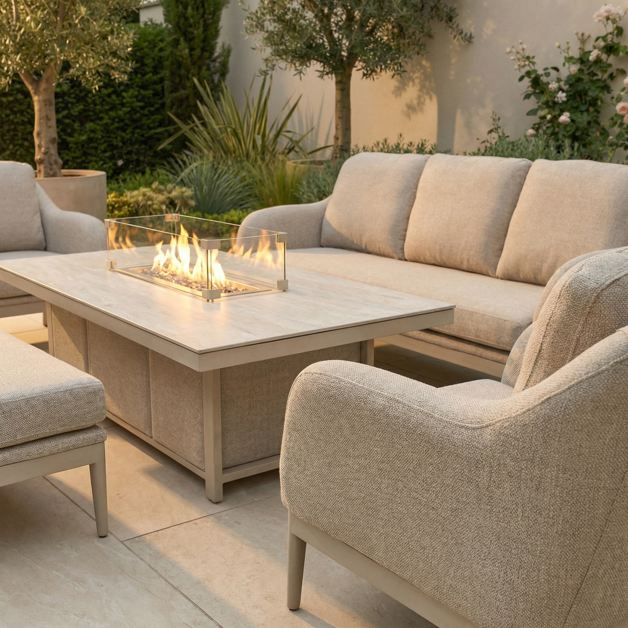 The Luna 3 Seat Outdoor Fabric Sofa Set with Rising Firepit Table in Fawn sits on the patio, offering cozy seating surrounded by lush green plants for a serene and inviting outdoor space.