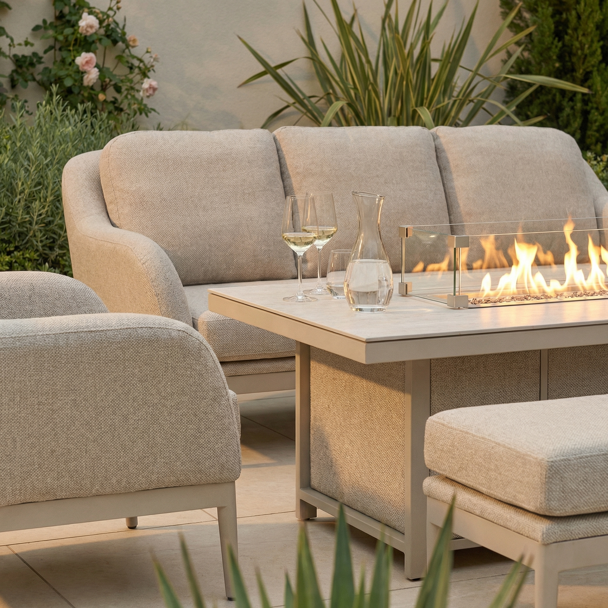 The Luna 3 Seat Outdoor Fabric Sofa Set with Rising Firepit Table in Fawn features beige, all-weather cushions and surrounds a firepit table, complemented by wine glasses and a carafe.