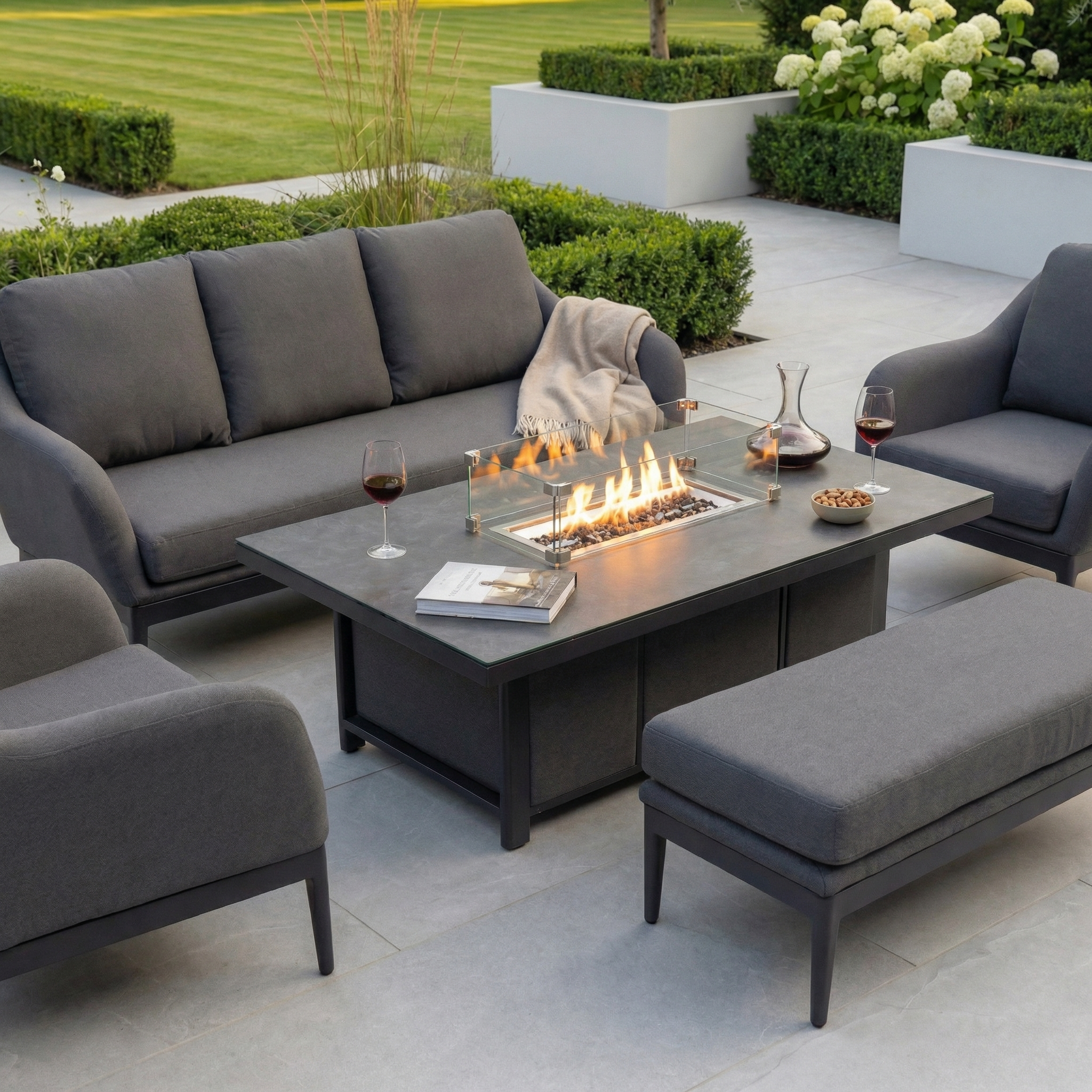A modern patio showcases the Luna 3 Seat Outdoor Fabric Sofa Set with Rising Firepit Table in Grey, accented by wine glasses and a landscaped garden.
