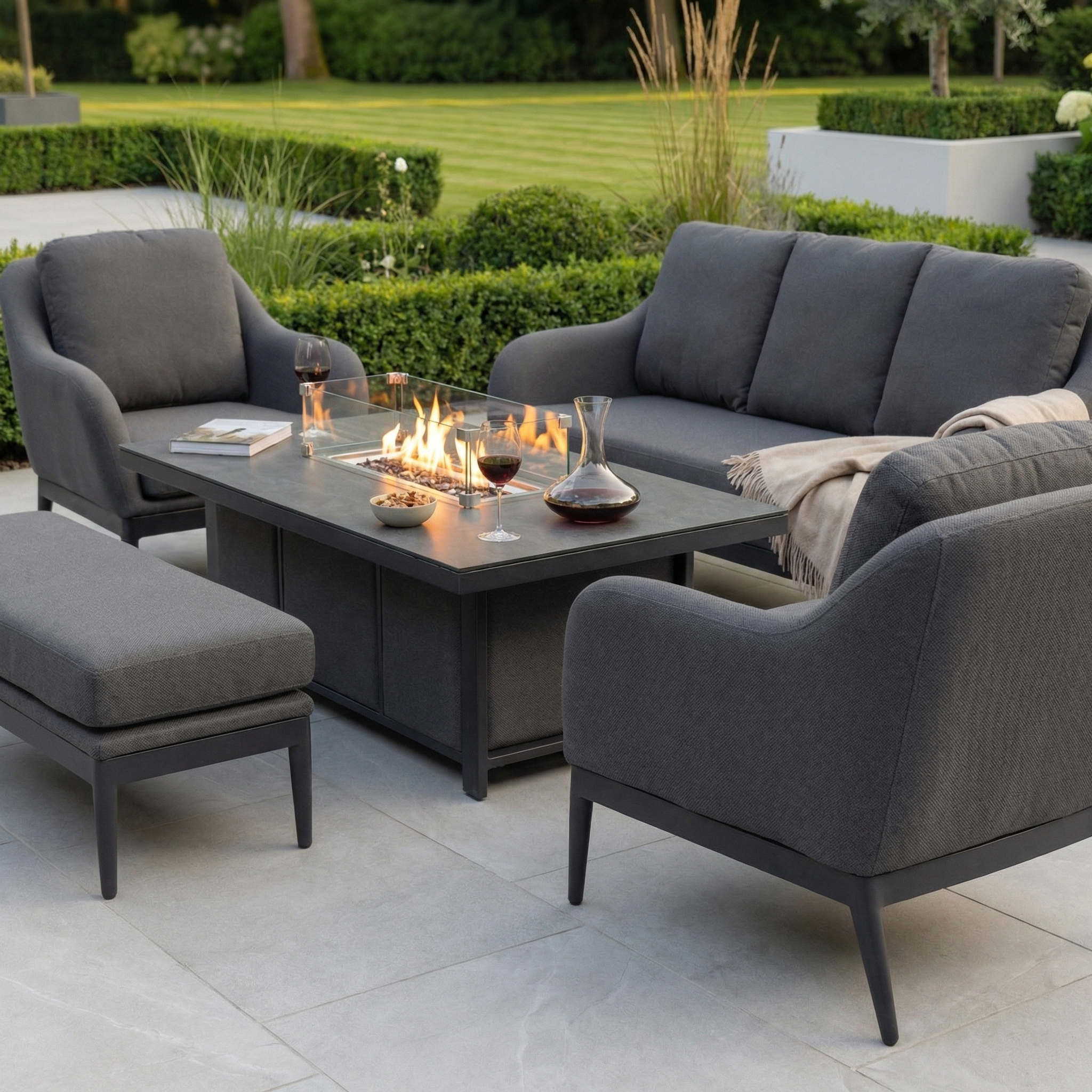 The Luna 3 Seat Outdoor Fabric Sofa Set with Rising Firepit Table in Grey features stylish seating and a sleek firepit table, perfect for outdoor gatherings amid greenery.