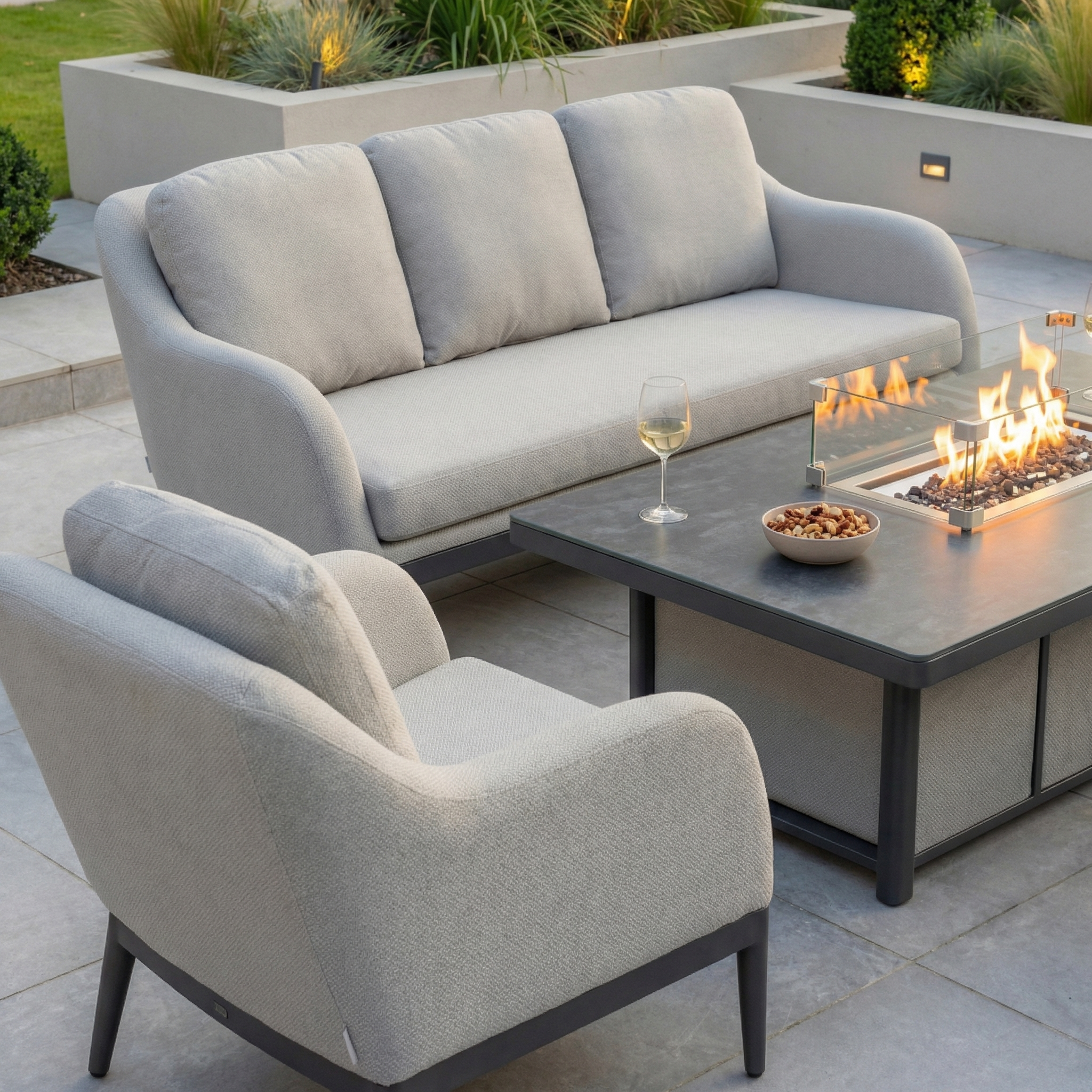 Outdoor patio featuring the Luna 3 Seat Outdoor Fabric Sofa Set with Rising Firepit Table in Oyster Grey, plus a wine glass and bowl of nuts on tiled flooring.