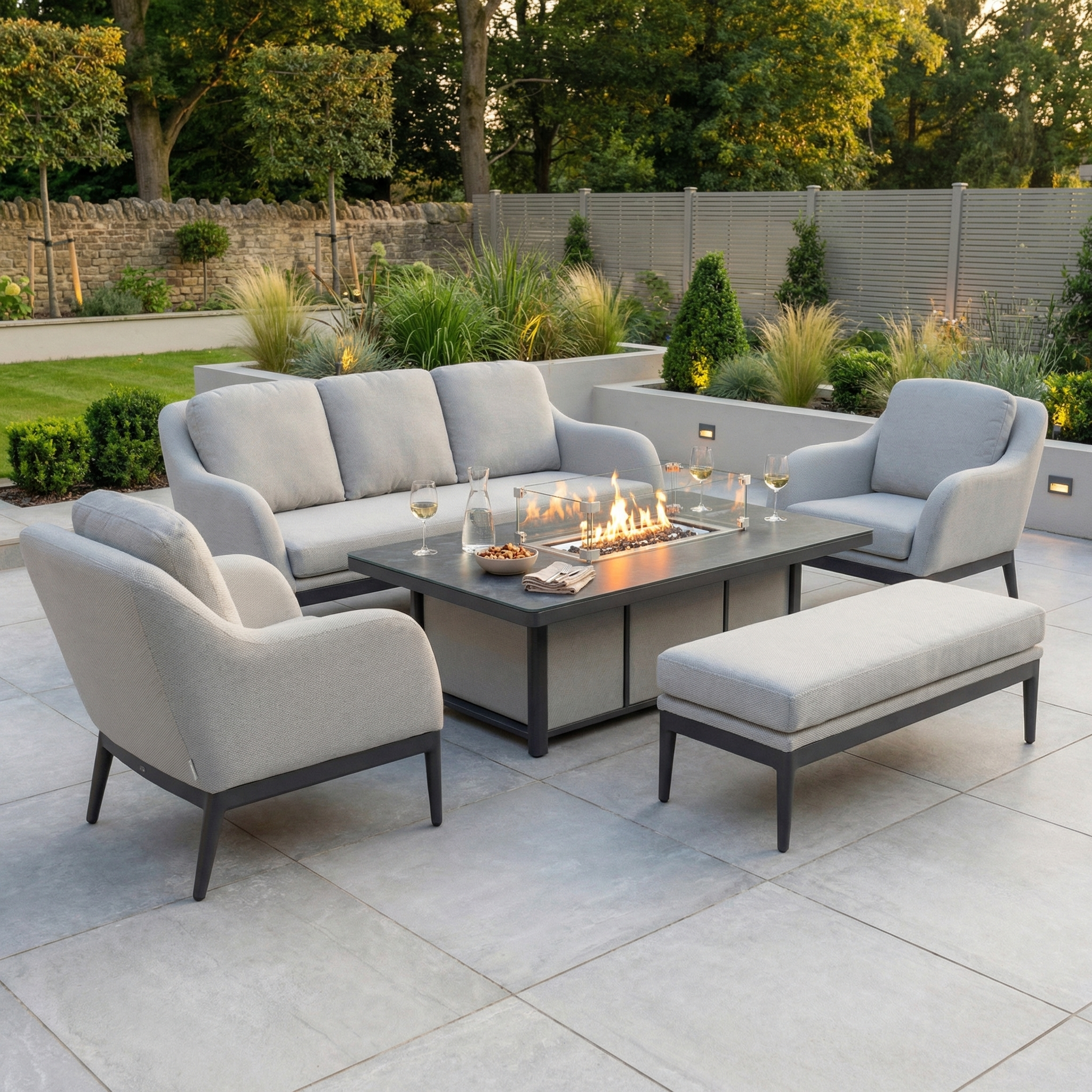 Enhance your patio with the Luna 3 Seat Outdoor Fabric Sofa Set in Oyster Grey, featuring cozy cushioned seating and a rising firepit table—all set amidst lush greenery for a modern outdoor retreat.