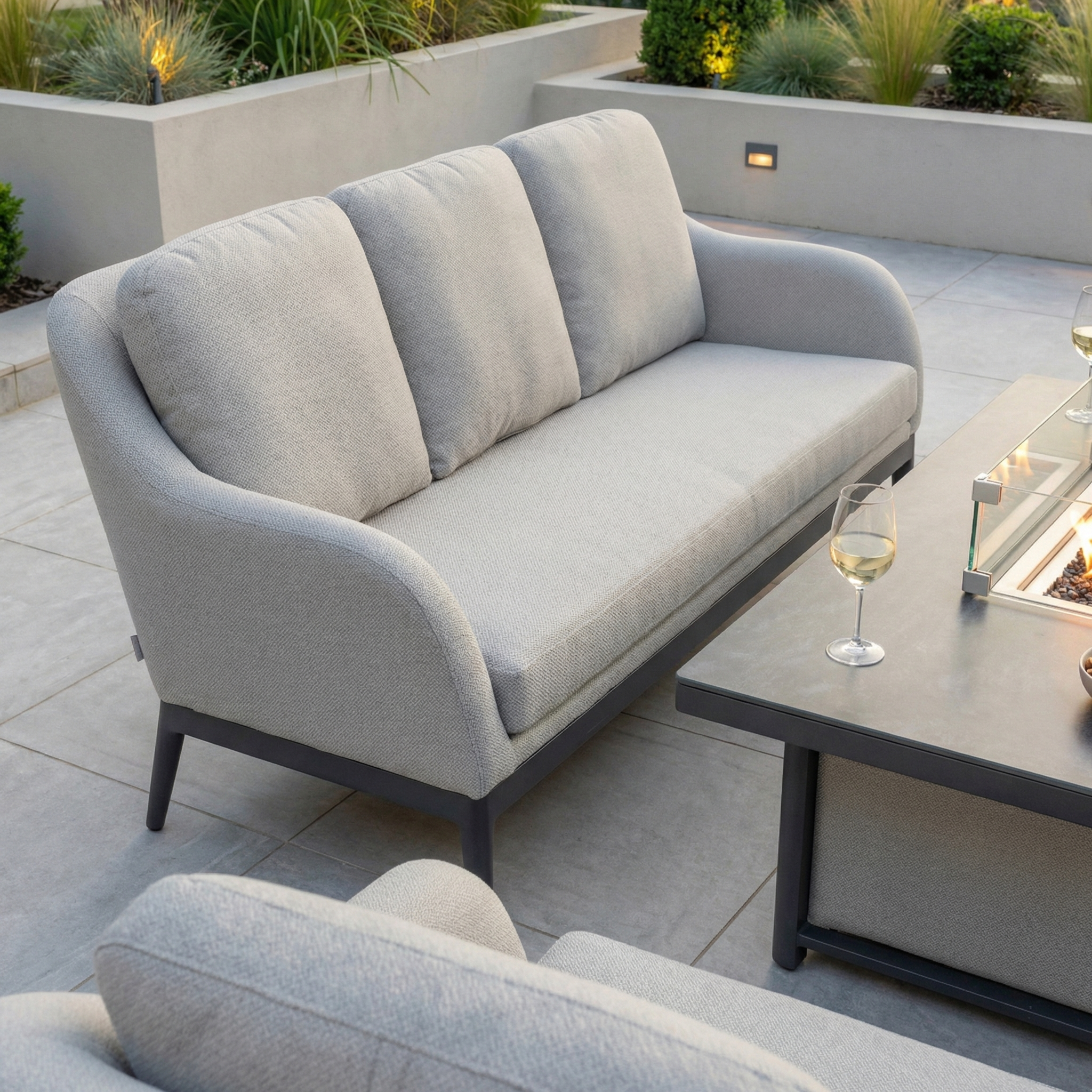 The Luna 3 Seat Outdoor Fabric Sofa Set with Rising Firepit Table in Oyster Grey sits on patio tiles with a glass of white wine, surrounded by modern landscaping.