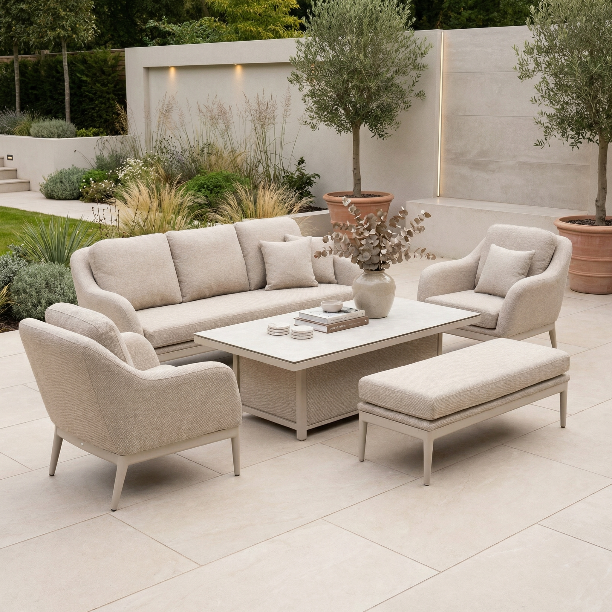 The Luna 3 Seat Outdoor Fabric Sofa Set with Rising Table in Fawn includes a sofa, chairs, and bench around a table, perfect for outdoor settings surrounded by lush plants and trees.