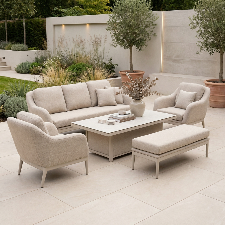 The Luna 3 Seat Outdoor Fabric Sofa Set with Rising Table in Fawn includes a sofa, chairs, and bench around a table, perfect for outdoor settings surrounded by lush plants and trees.