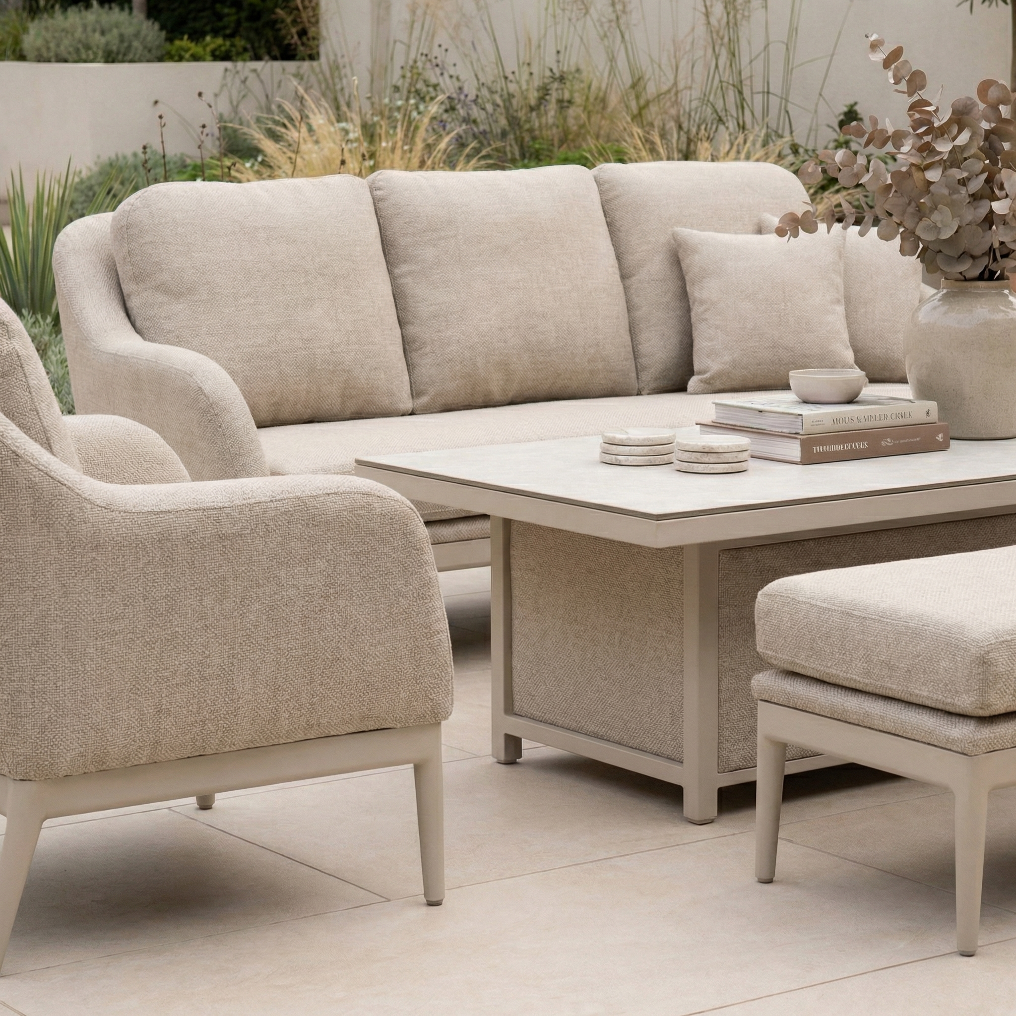 The Luna 3 Seat Outdoor Fabric Sofa Set with Rising Table in Fawn features a sofa, chair, and table arranged on a tiled floor. Accessories include books and a vase with dried leaves, creating an inviting outdoor setting.