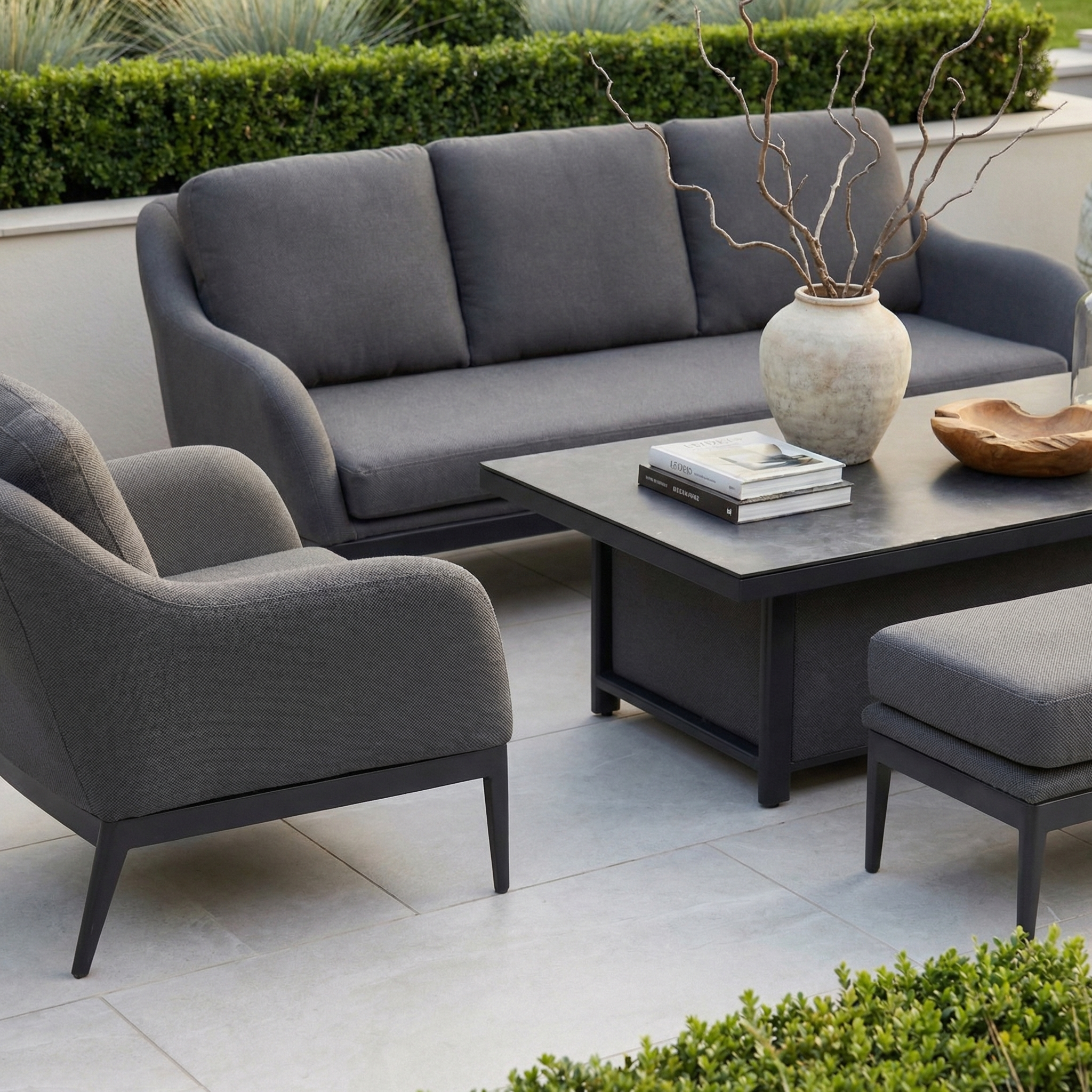 The Luna 3 Seat Outdoor Fabric Sofa Set with Rising Table in Grey features stain-resistant, all-weather design and includes a sofa, armchair, and table accented by a decorative vase and books.