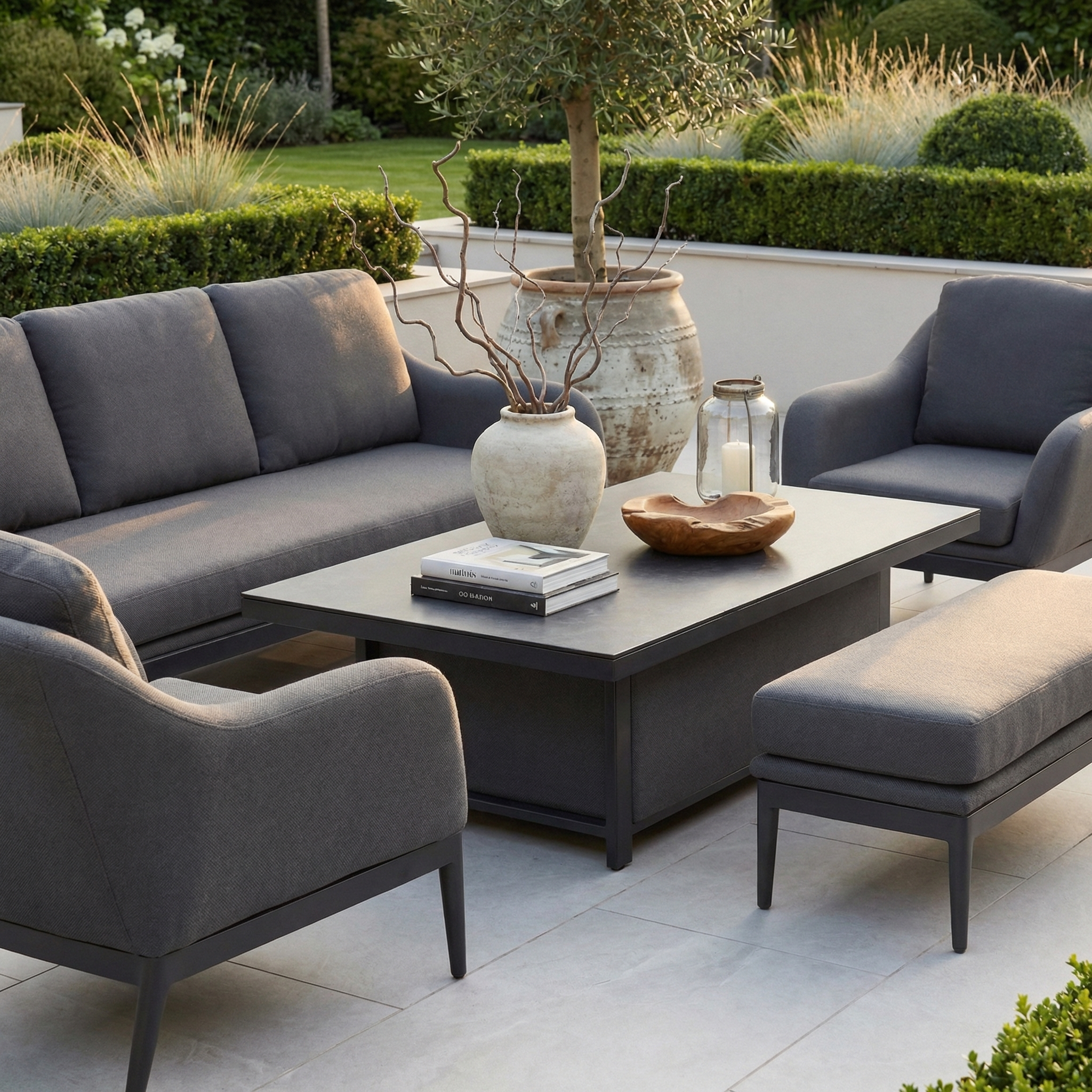 Modern patio featuring the Luna 3 Seat Outdoor Fabric Sofa Set with Rising Table in Grey, stain-proof seating, books and a vase on the table, garden decor, and lush greenery all around.