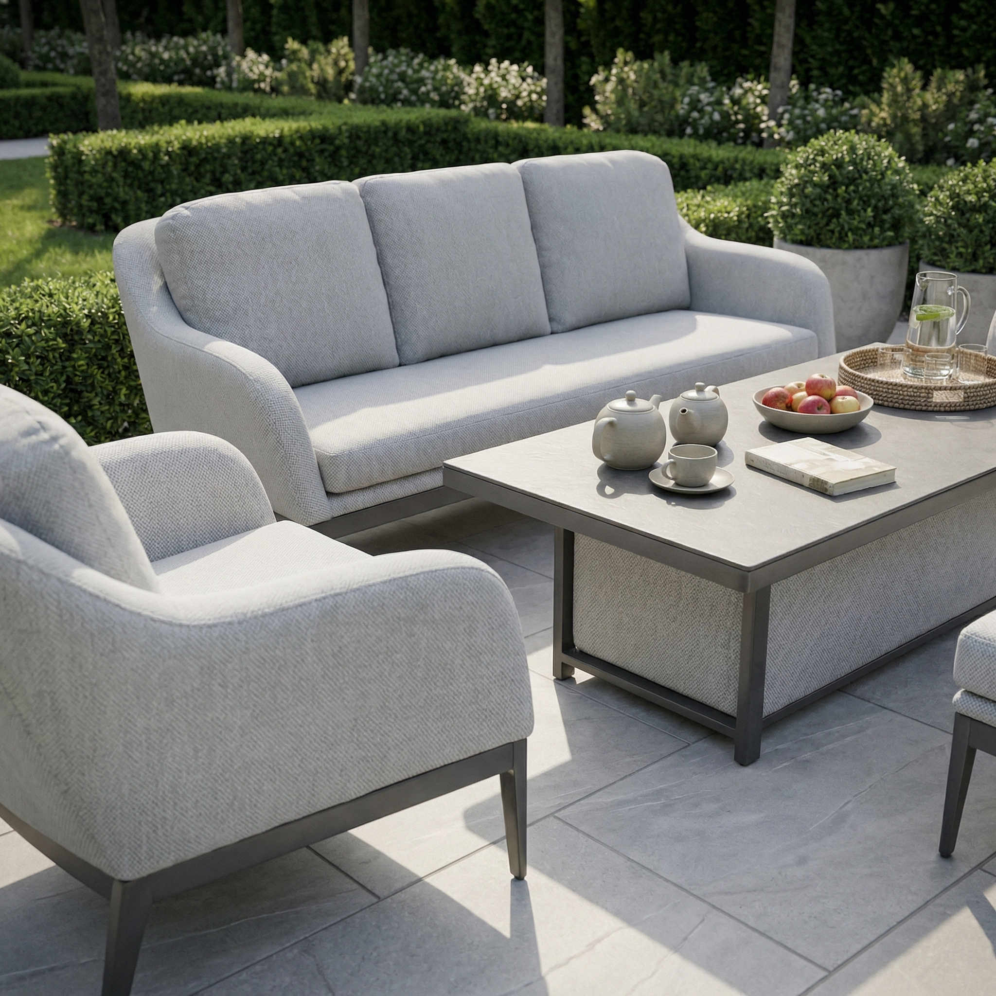 The Luna 3 Seat Outdoor Fabric Sofa Set with Rising Table in Oyster Grey offers a maintenance-free retreat, including a table, tea set, fruit plate, and tray—perfect for stylish outdoor living amid lush greenery.