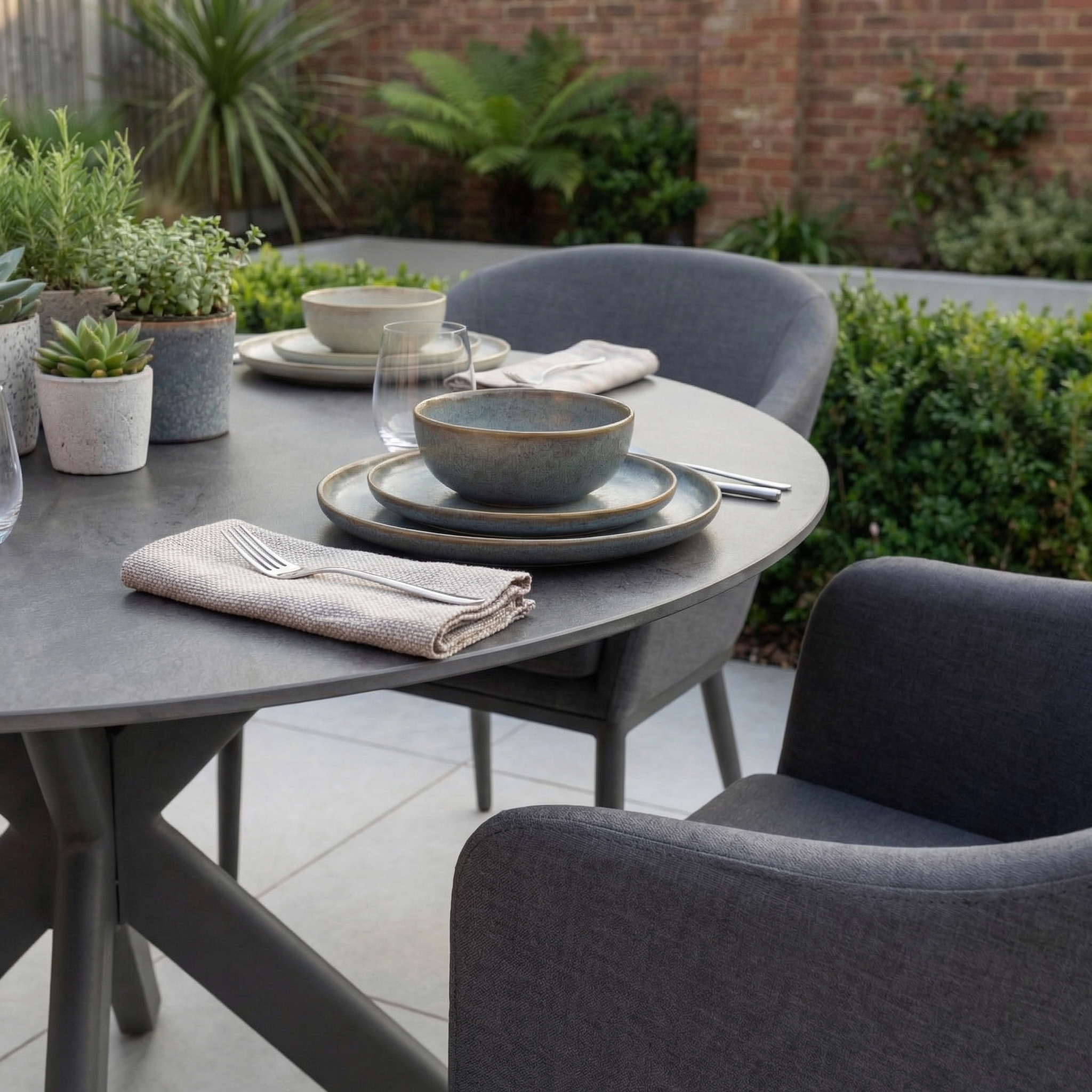 The Luna 4 Seat Round Outdoor Fabric Dining Set with Albatross Table in Grey is shown with stylish chairs, tableware, and décor, set against a lush green backdrop.