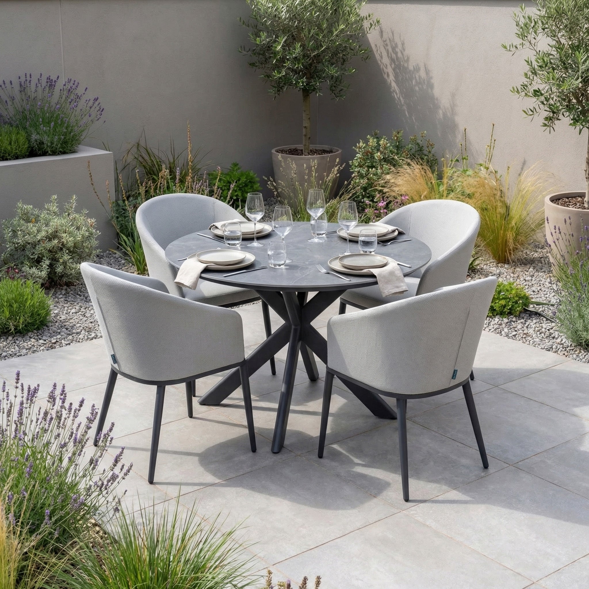 The Luna 4 Seat Round Outdoor Fabric Dining Set with Albatross Table in Oyster Grey, set with plates and glasses, invites al fresco dining among lush plants and gravel.