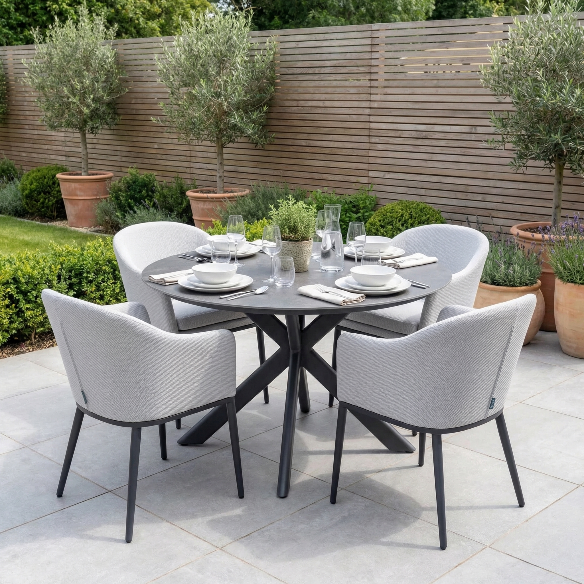 The Luna 4 Seat Round Outdoor Fabric Dining Set with Albatross Table in Oyster Grey provides stylish al fresco dining for four, set amidst olive trees and potted plants on the patio.
