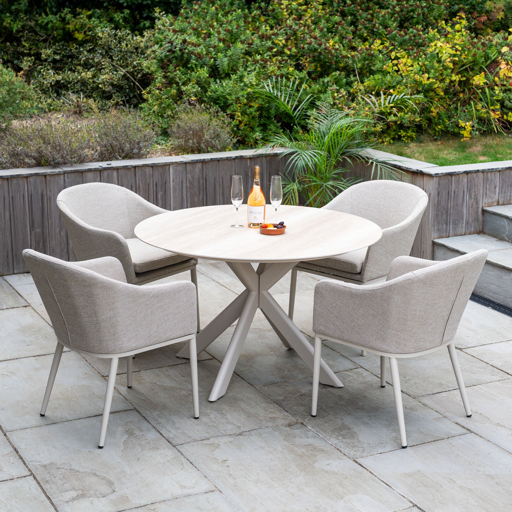 The Luna 4 Seat Round Outdoor Fabric Dining Set with Albatross Table in Fawn features a ceramic table and four beige chairs, perfect for patios. Enjoy drinks and snacks outdoors, surrounded by lush greenery with this all-weather set.