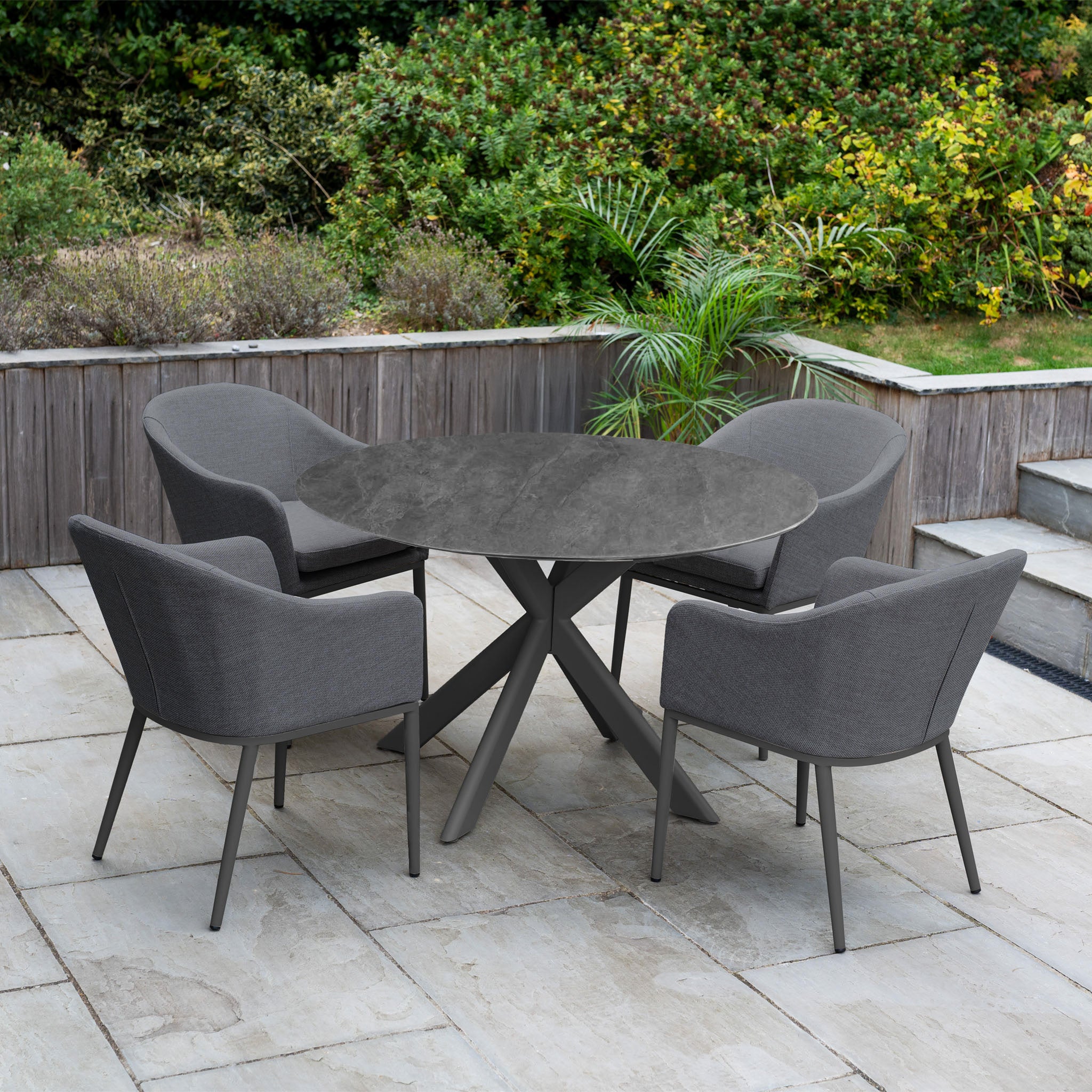 The Luna 4 Seat Round Outdoor Fabric Dining Set with Albatross Table in Grey is arranged on a stone patio, surrounded by lush greenery.