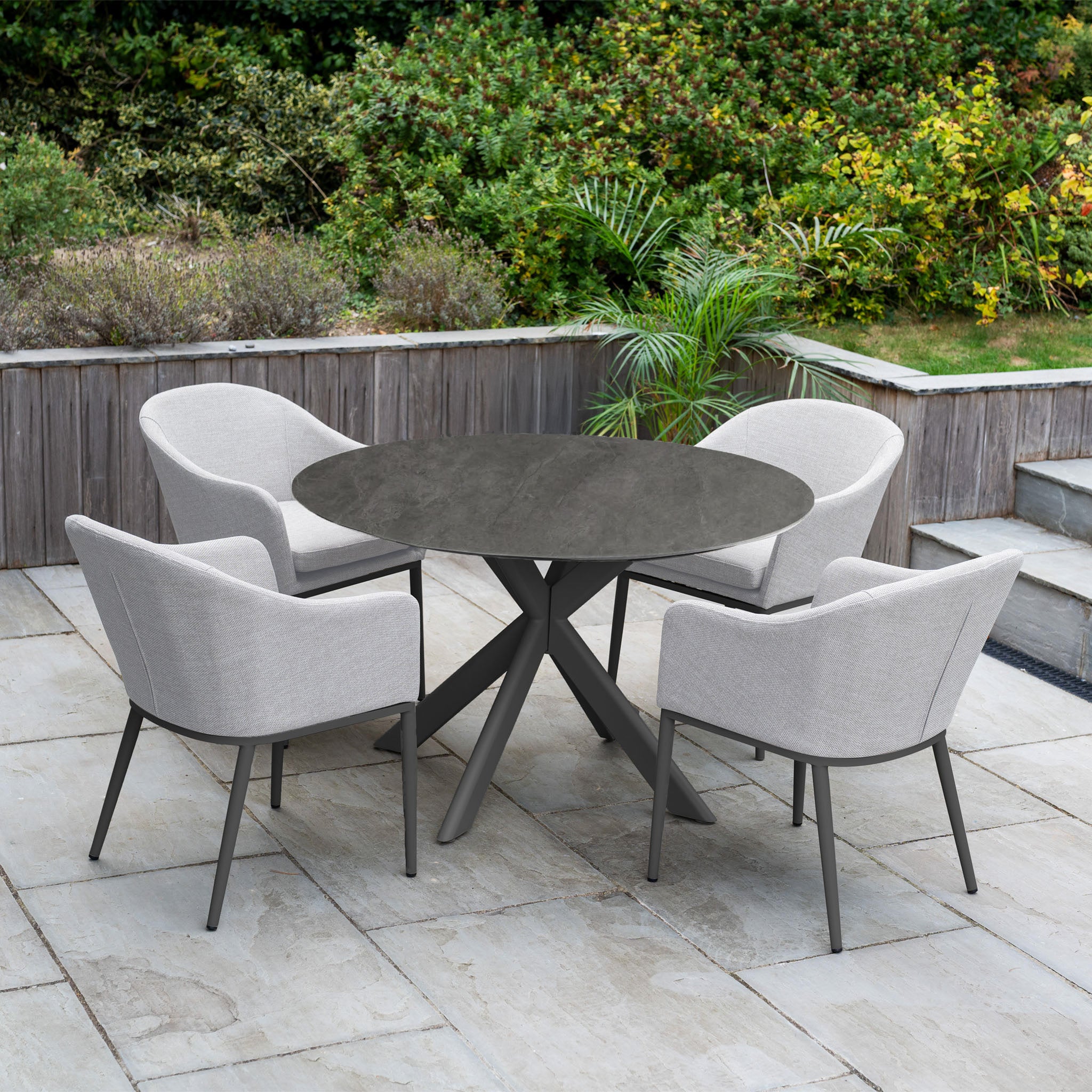 The Luna 4 Seat Round Outdoor Fabric Dining Set with Albatross Table in Oyster Grey sits on a stone patio surrounded by greenery—ideal for stylish al fresco dining.