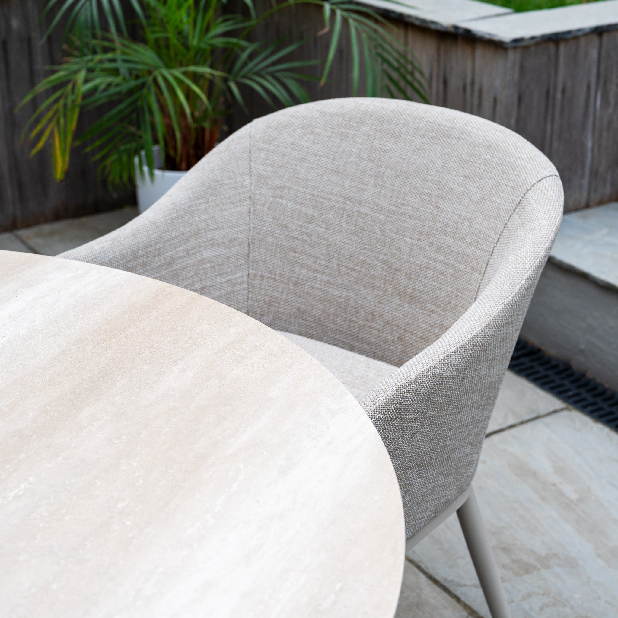 The Luna 4 Seat Round Outdoor Fabric Dining Set with Albatross Table in Fawn is arranged on the patio, with its beige chairs beside a round ceramic table and a green plant in the background.