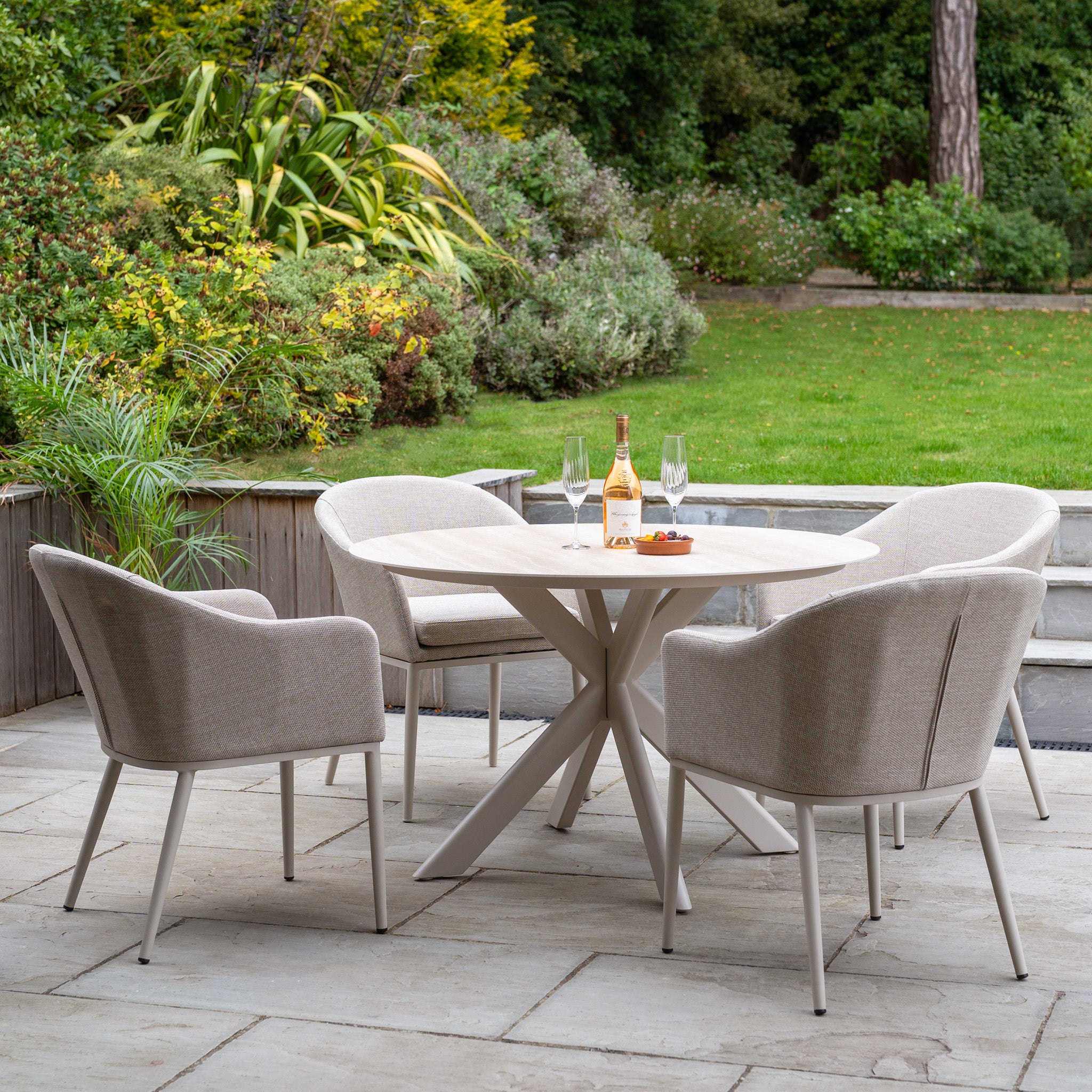The Luna 4 Seat Round Outdoor Fabric Dining Set with Albatross Table in Fawn creates an inviting spot for dining with wine and glasses on a stone patio surrounded by a lush garden.