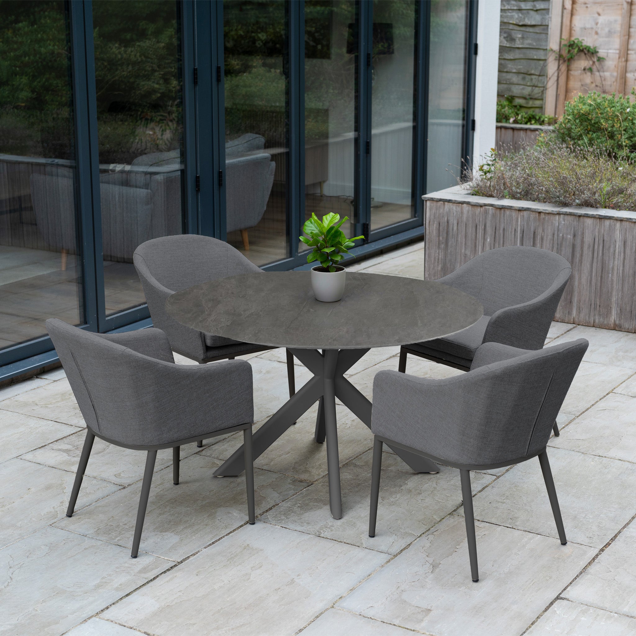 The Luna 4 Seat Round Outdoor Fabric Dining Set with Albatross Table in Grey, arranged on a tiled terrace near glass doors, includes four chairs and a round grey table, complemented by a potted plant for stylish outdoor dining.