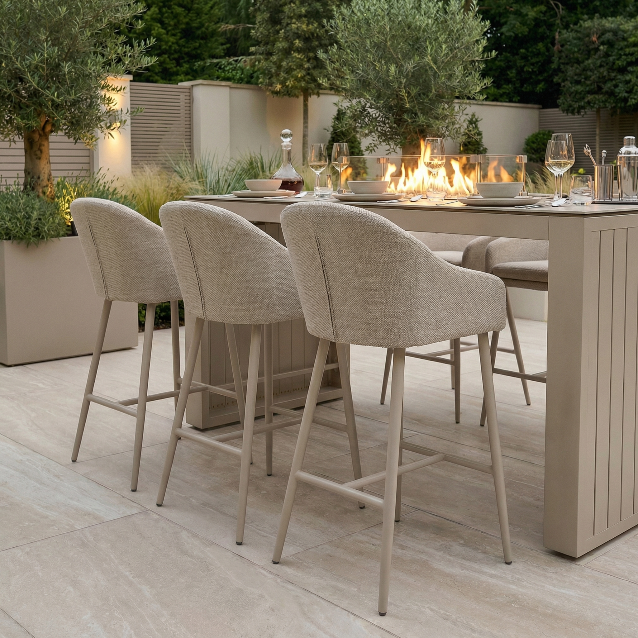 Outdoor dining area with the Luna 6 Seat Outdoor Fabric Ceramic Firepit Bar Set in Fawn, featuring beige bar stools, a tall table, wine glasses, and a central fire feature—all crafted from durable all-weather materials.