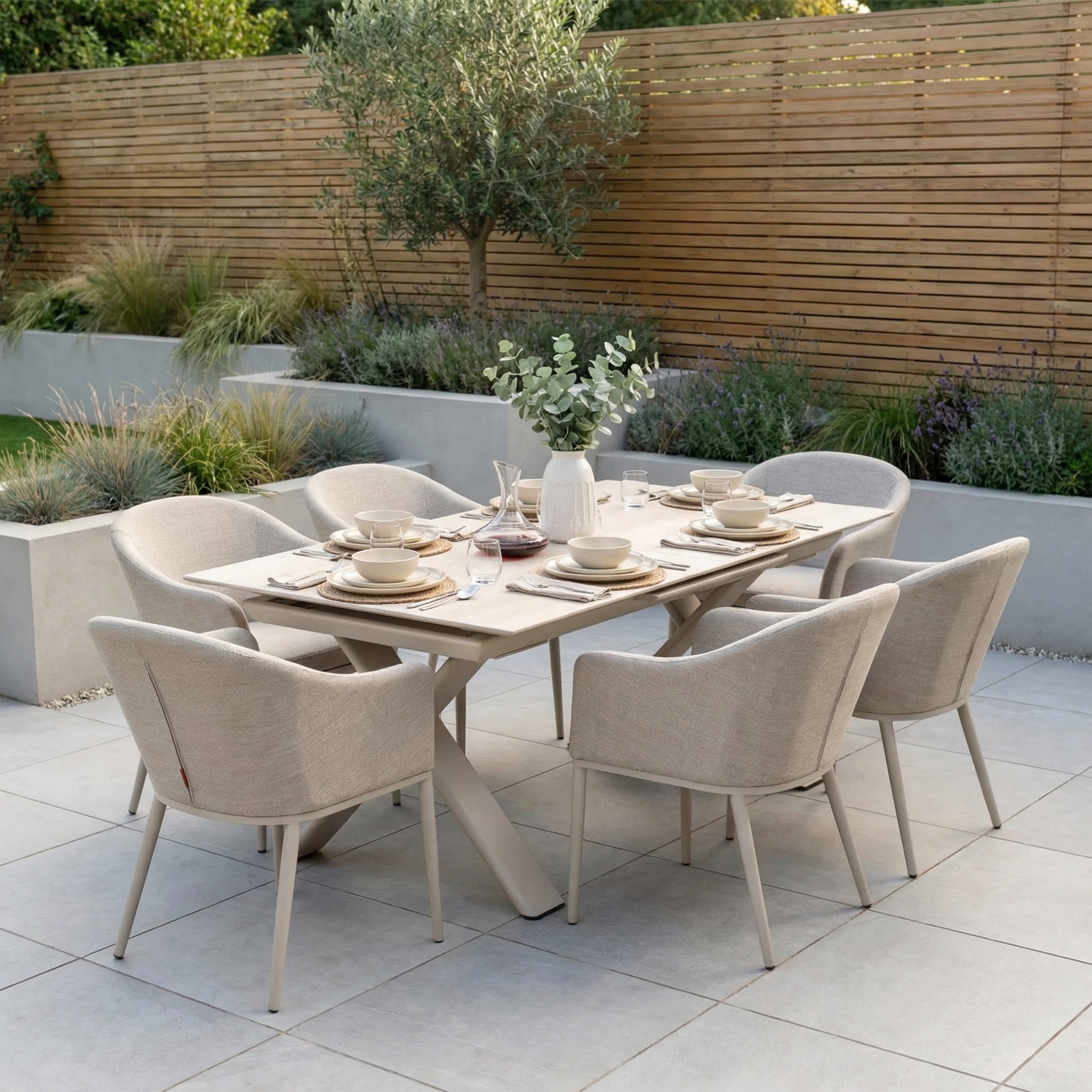 The Luna 6 Seat Outdoor Fabric Dining Set with Albatross Extending Table in Fawn is shown arranged for dining on a patio with six all-weather chairs, surrounded by lush plants and a wooden fence.