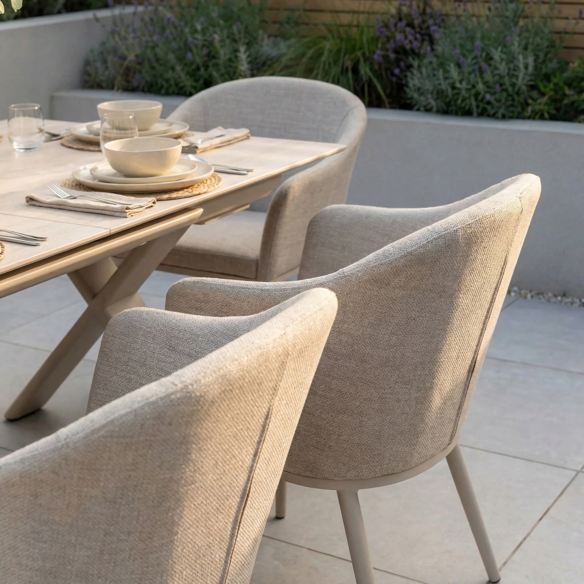 The Luna 6 Seat Outdoor Fabric Dining Set with Albatross Extending Table in Fawn is arranged on a patio with lush plants, featuring all-weather dining chairs and neutral tableware for modern outdoor dining.