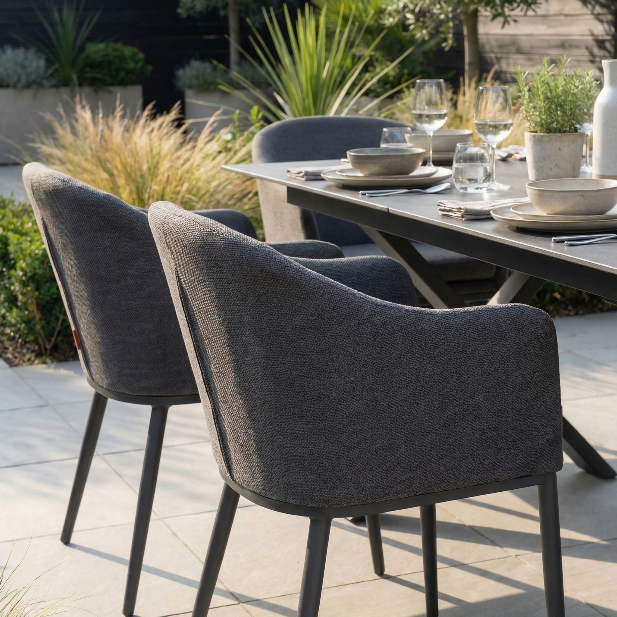 The Luna 6 Seat Outdoor Fabric Dining Set with Albatross Extending Table in Grey features cushioned chairs arranged around a table set with plates and glasses, complemented by nearby plants.