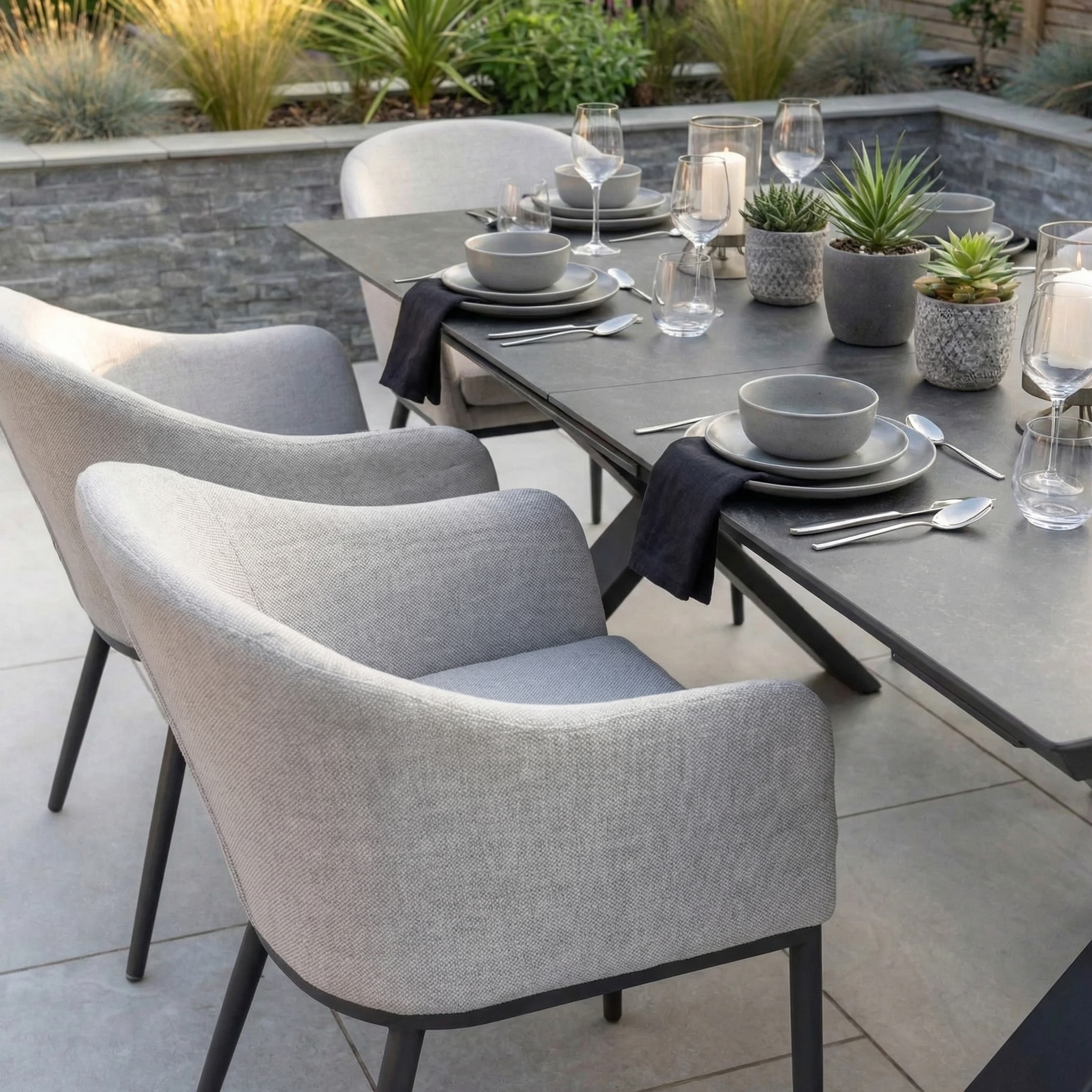 The Luna 6 Seat Outdoor Fabric Dining Set with Albatross Extending Table in Oyster Grey features grey chairs, an extending table, and stylish décor including place settings, candles, and succulent plants.