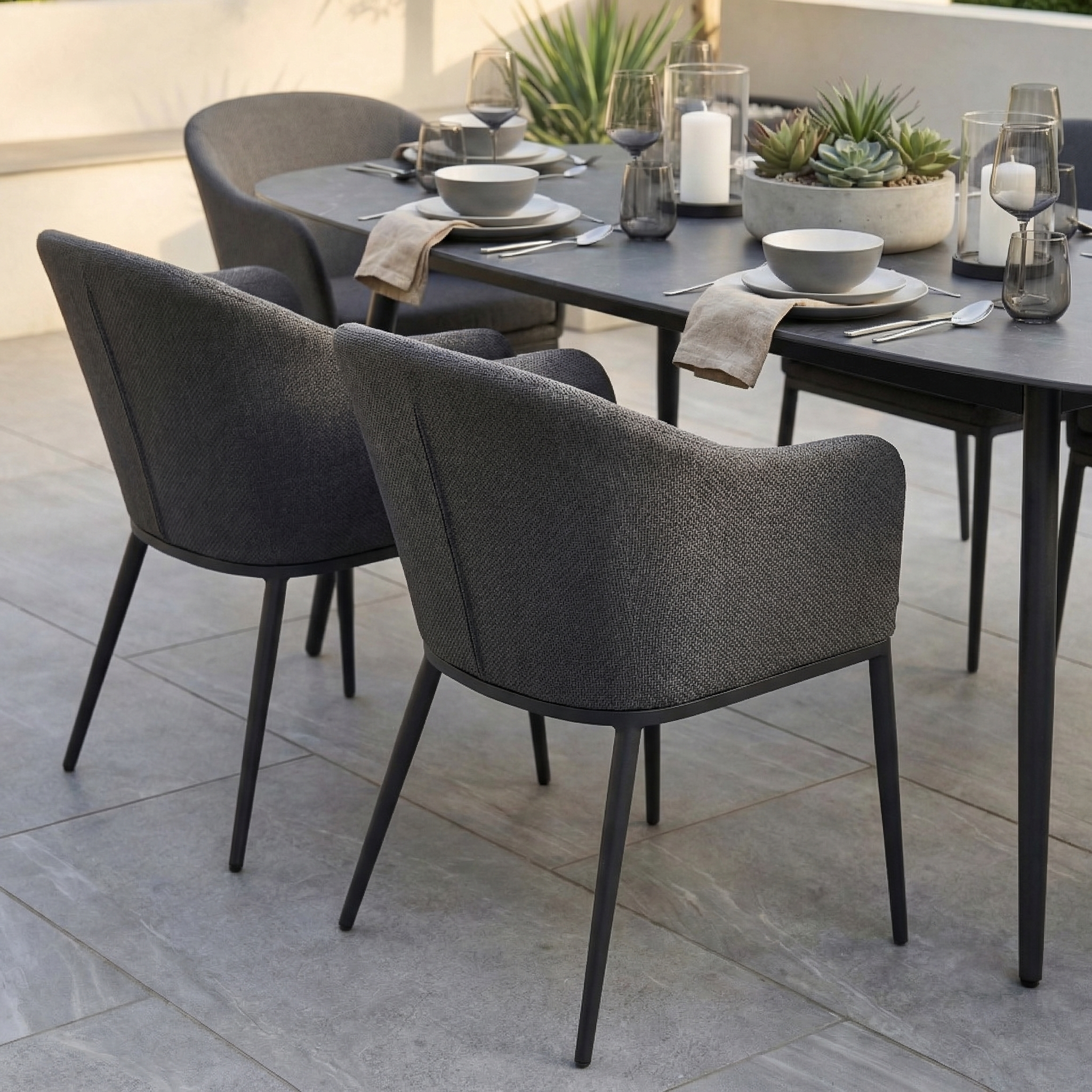 The Luna 6 Seat Outdoor Fabric Oval Ceramic Dining Set in Grey features stain-resistant grey upholstered chairs, and comes styled with dishes, candles, and a succulent centerpiece for modern all-weather dining.