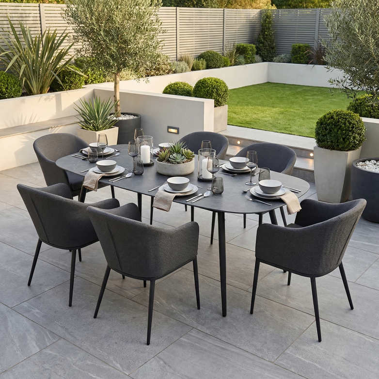 The Luna 6 Seat Outdoor Fabric Oval Ceramic Dining Set in Grey features six dark, stain-resistant chairs, arranged with plates, glasses, and candles in a landscaped garden.