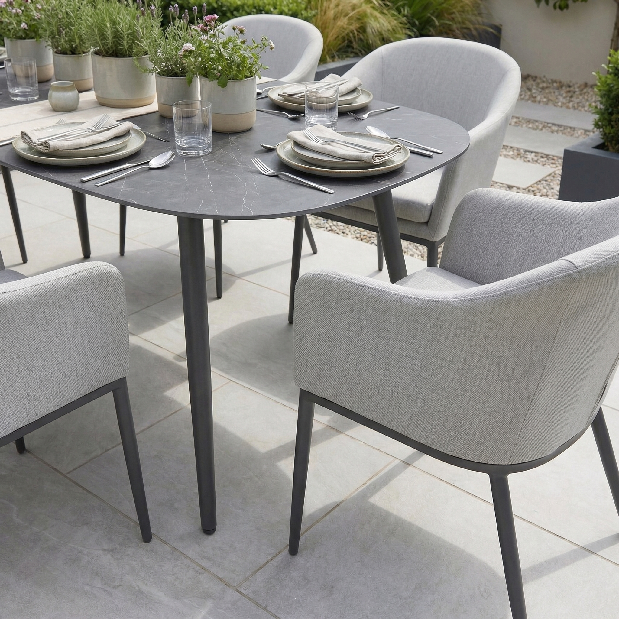 Luna 6 Seat Outdoor Fabric Oval Ceramic Dining Set in Oyster Grey features cushioned chairs and durable all-weather materials, styled with plates, glasses, and potted plants.