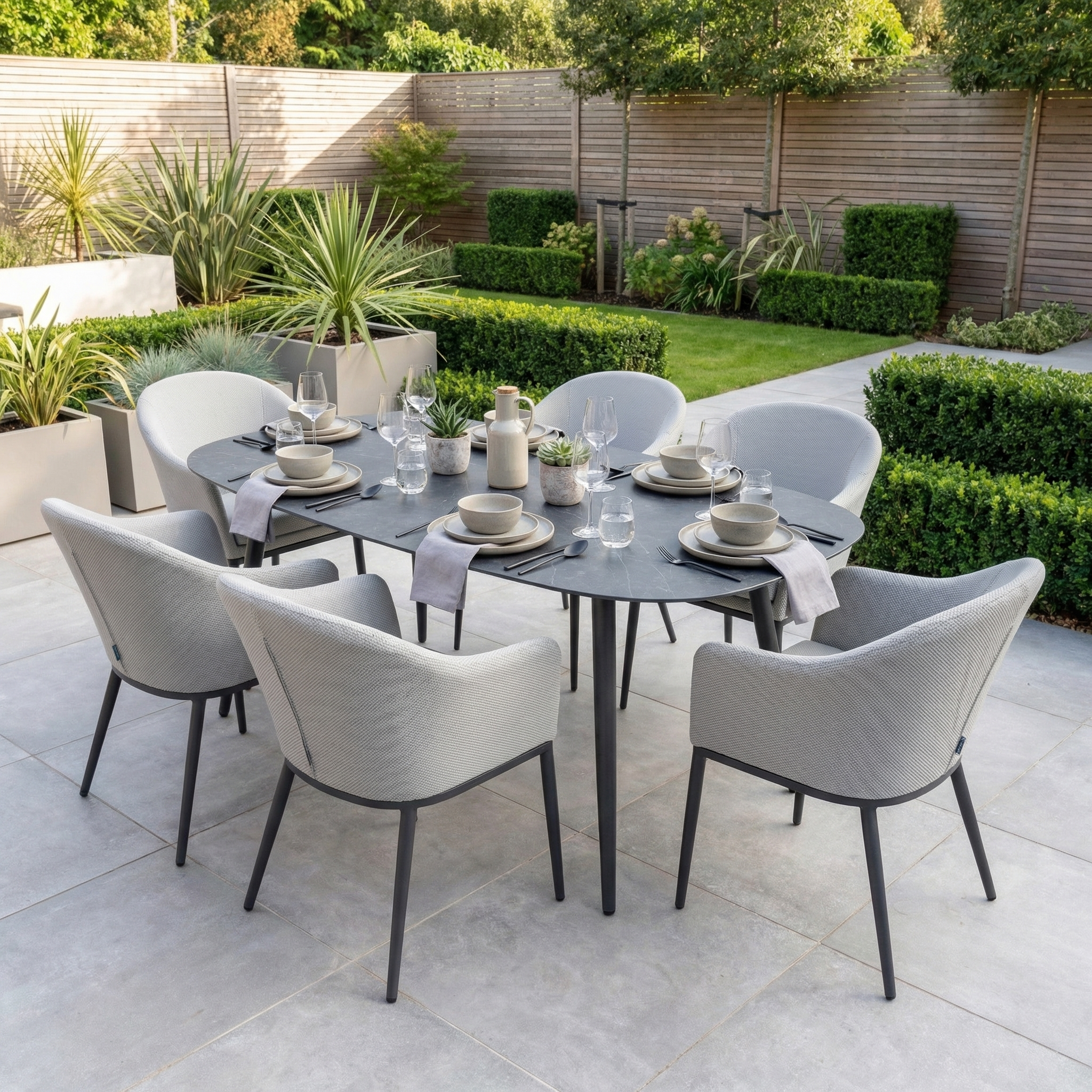 The Luna 6 Seat Outdoor Fabric Oval Ceramic Dining Set in Oyster Grey features all-weather materials, ideal for al fresco dining on your patio amid lush greenery.