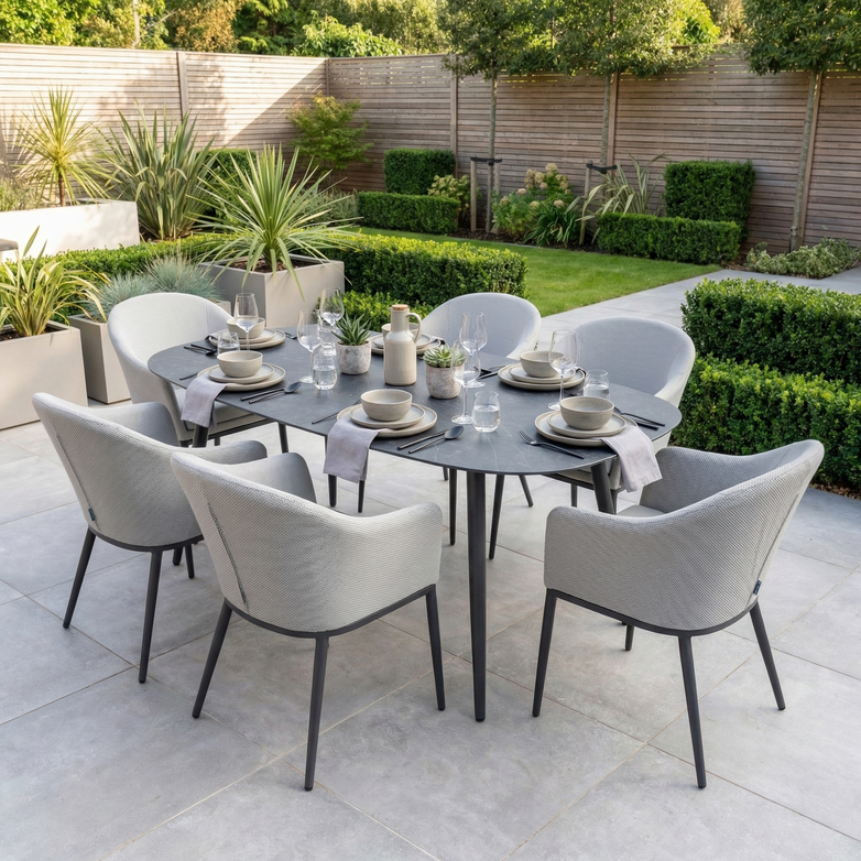 The Luna 6 Seat Outdoor Fabric Oval Ceramic Dining Set in Oyster Grey features all-weather materials, ideal for al fresco dining on your patio amid lush greenery.