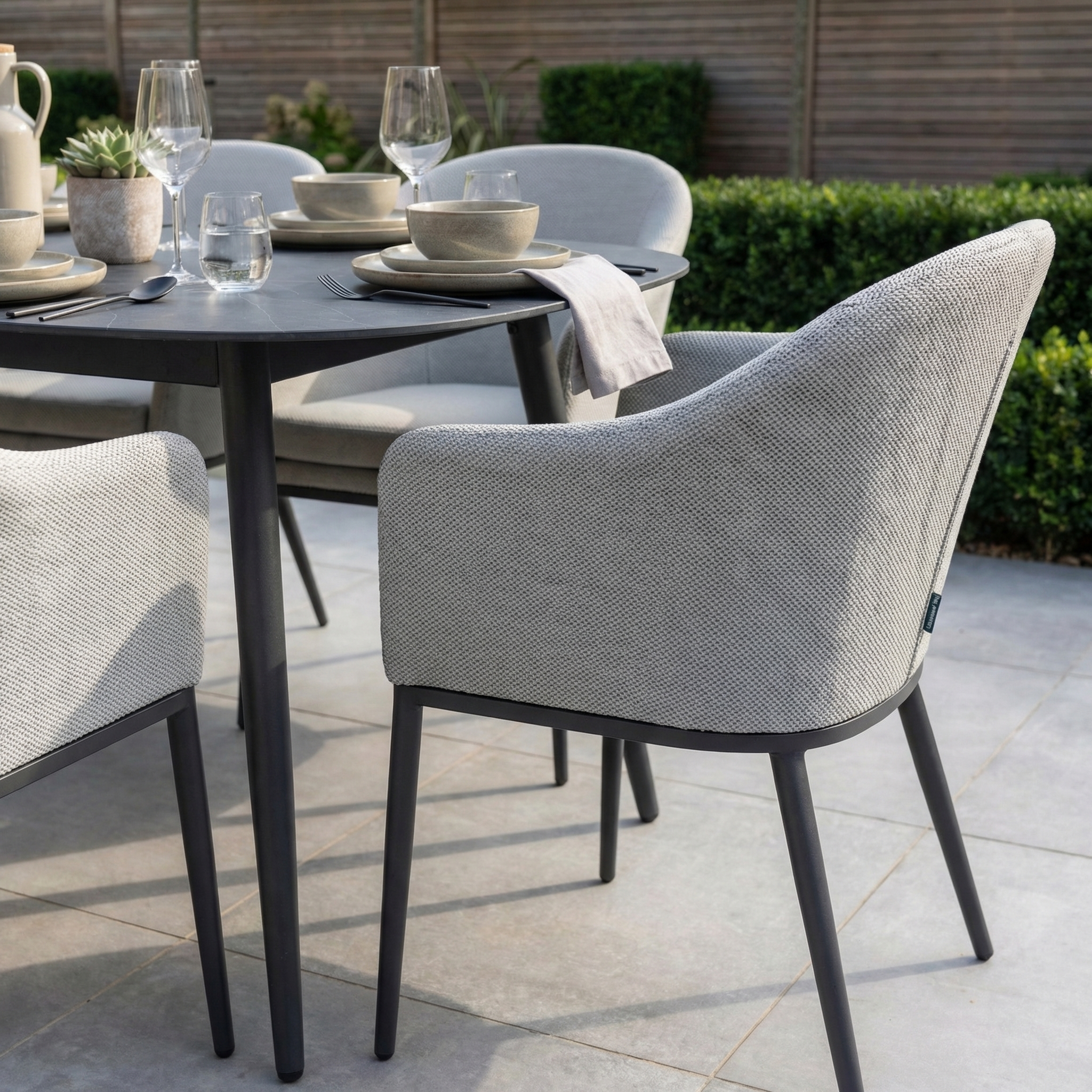 The Luna 6 Seat Outdoor Fabric Oval Ceramic Dining Set in Oyster Grey features gray chairs and all-weather materials, set with dishes and glassware on a tiled patio for lasting outdoor style.
