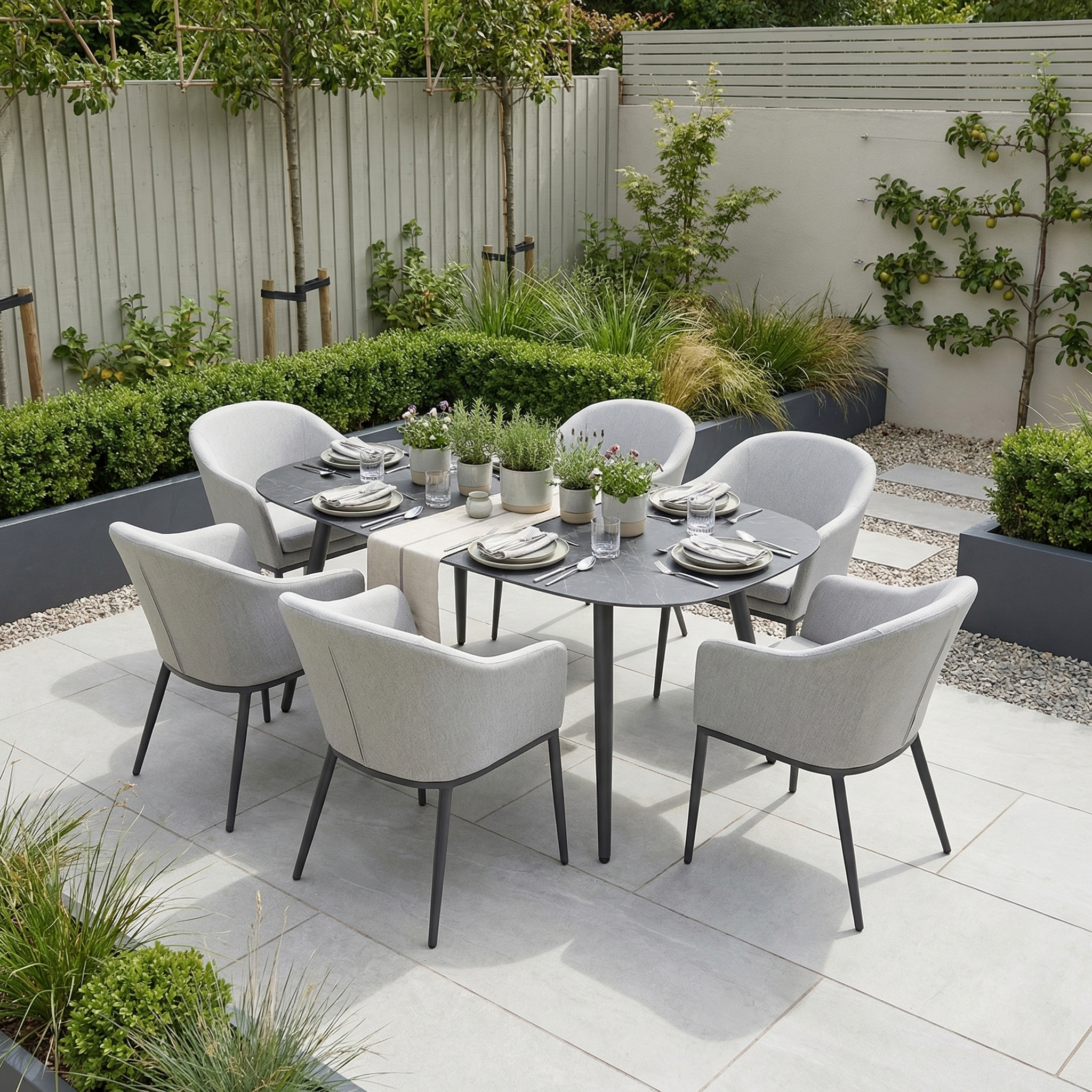 The Luna 6 Seat Outdoor Fabric Oval Ceramic Dining Set in Oyster Grey features durable, all-weather materials and stylish grey chairs, perfect for outdoor spaces with lush plants and a wooden fence backdrop.
