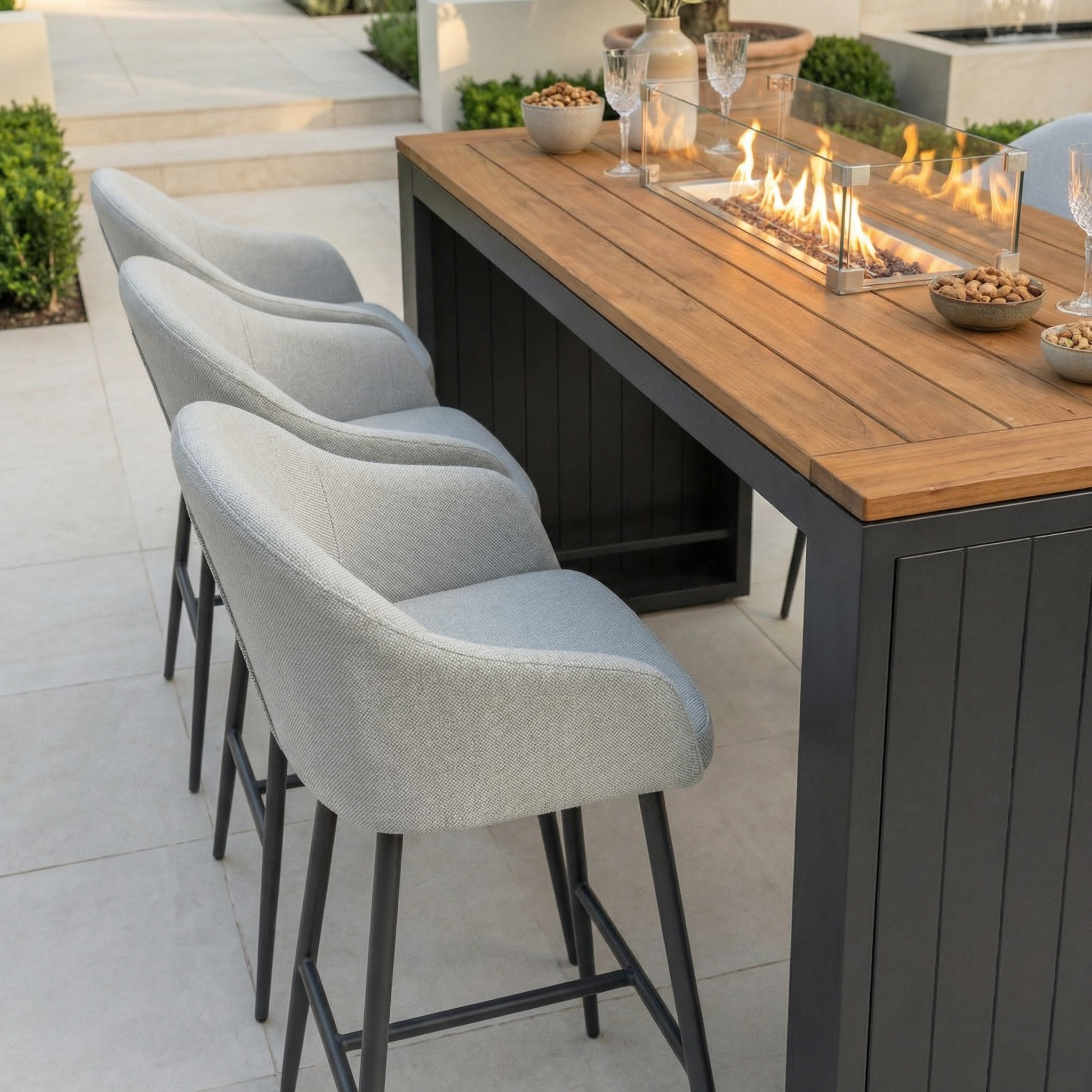 The Luna 6 Seat Outdoor Fabric Teak Firepit Bar Set in Oyster Grey features teak construction, a built-in fire feature, and six cushioned bar stools with all-weather Luna fabric for stylish outdoor entertaining.