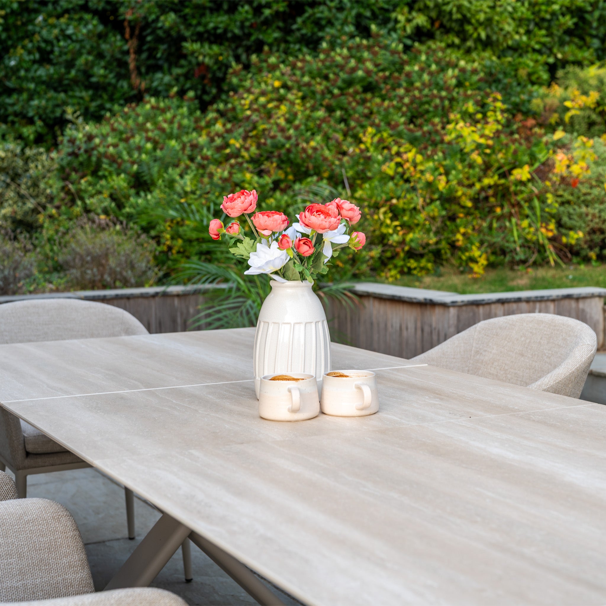A vase of pink flowers and two mugs rest on the Luna 6 Seat Outdoor Fabric Dining Set with Albatross Extending Table in Fawn, surrounded by elegant chairs and lush greenery.