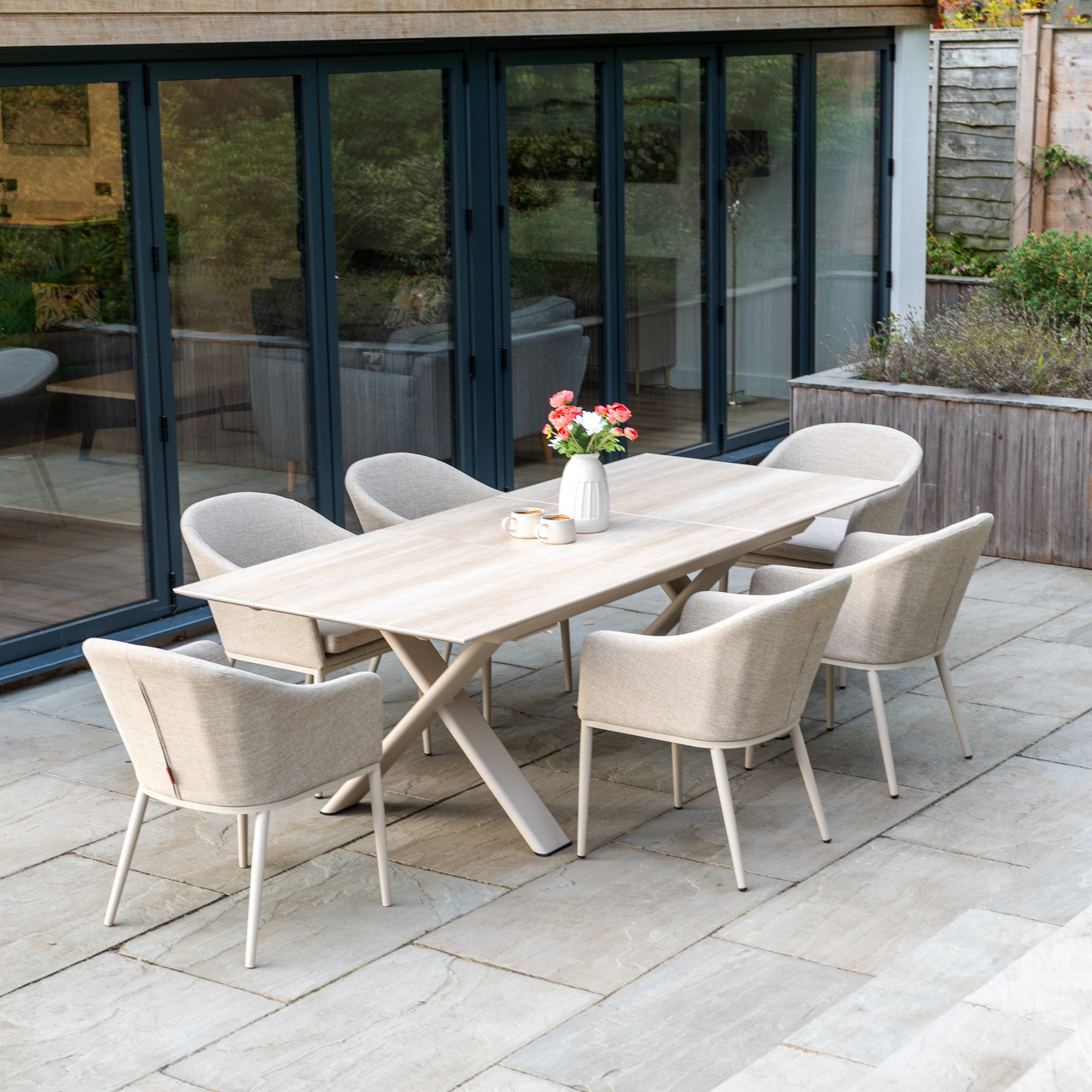 The Luna 6 Seat Outdoor Fabric Dining Set with Albatross Extending Table in Fawn features six all-weather beige chairs and a table, styled here with a white vase holding pink flowers.