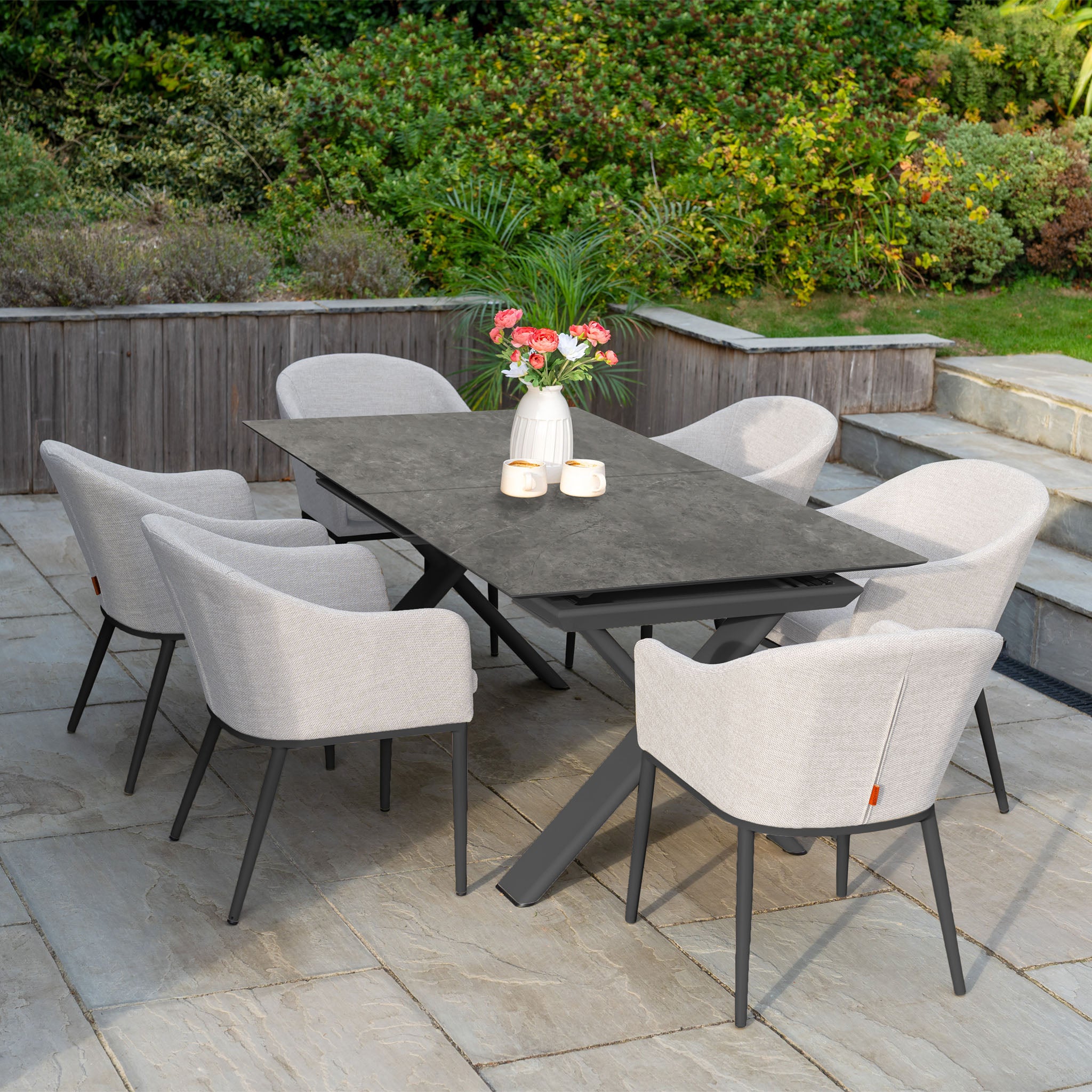 The Luna 6 Seat Outdoor Fabric Dining Set with Albatross Extending Table in Oyster Grey features six all-weather fabric chairs, ideal for modern patios surrounded by flowers and lush greenery.