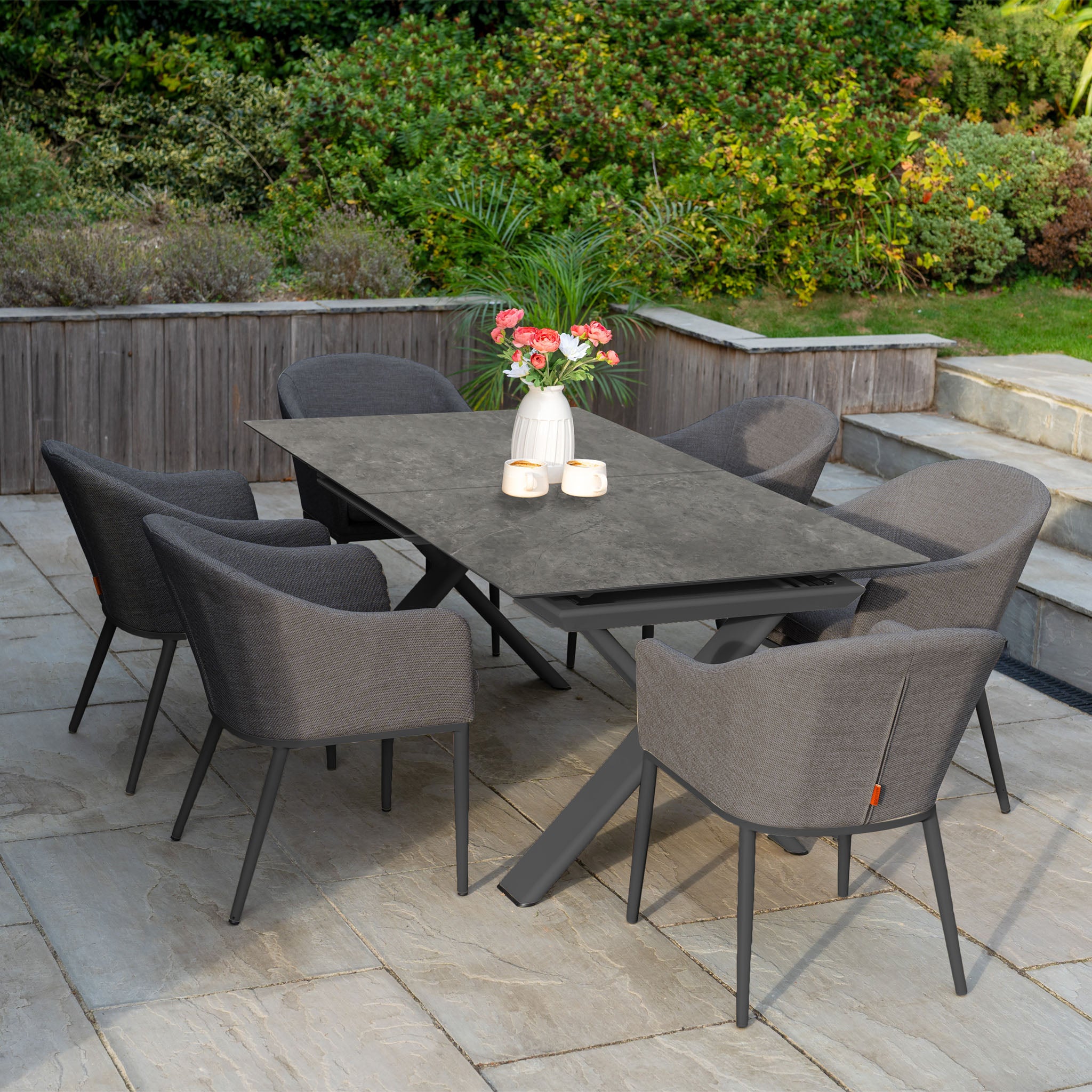 The Luna 6 Seat Outdoor Fabric Dining Set with Albatross Extending Table in Grey is arranged on a stone patio surrounded by greenery, featuring six chairs and a flower vase centerpiece.