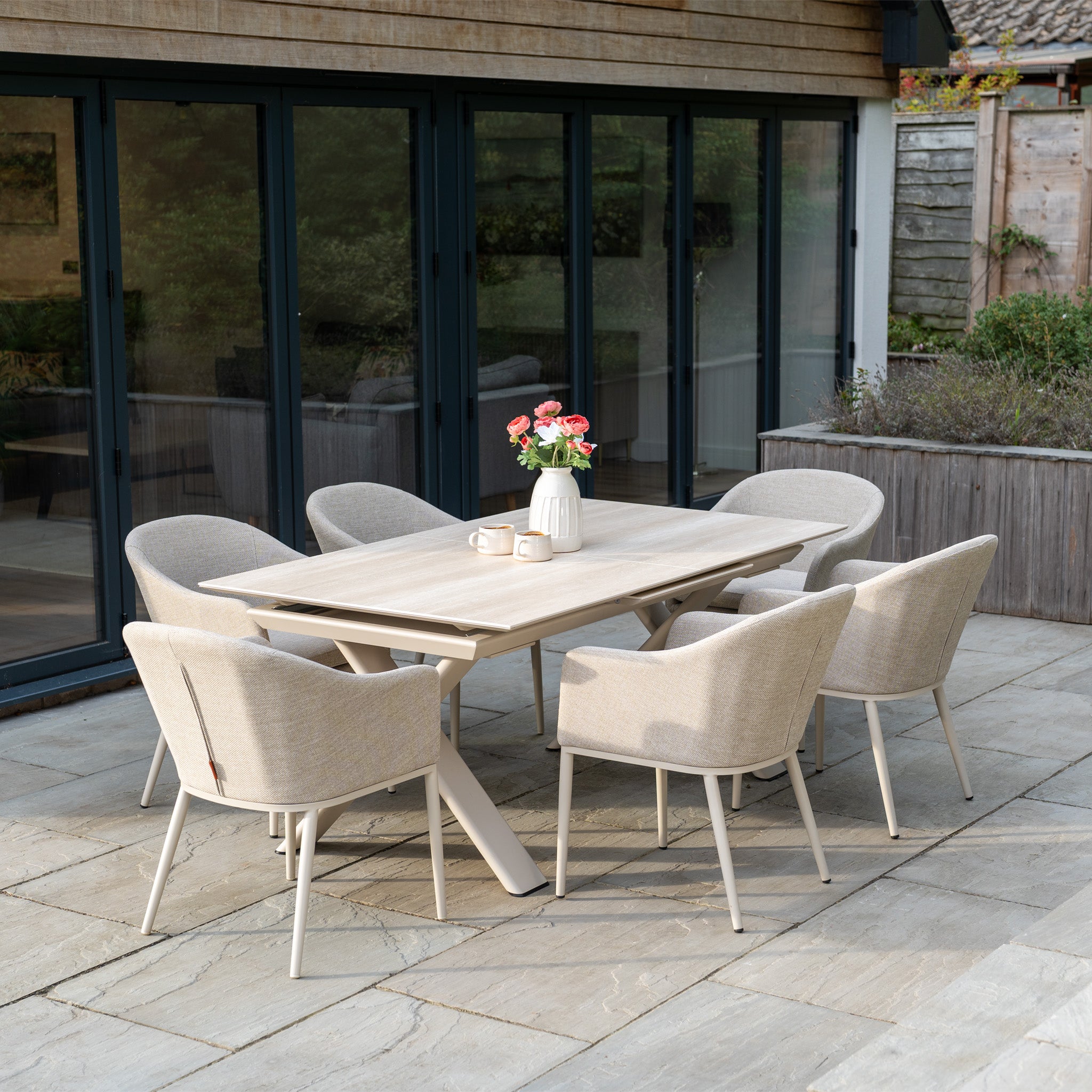 The Luna 6 Seat Outdoor Fabric Dining Set with Albatross Extending Table in Fawn features six all-weather chairs around a stylish table topped with a white vase of flowers, perfect for your stone patio.