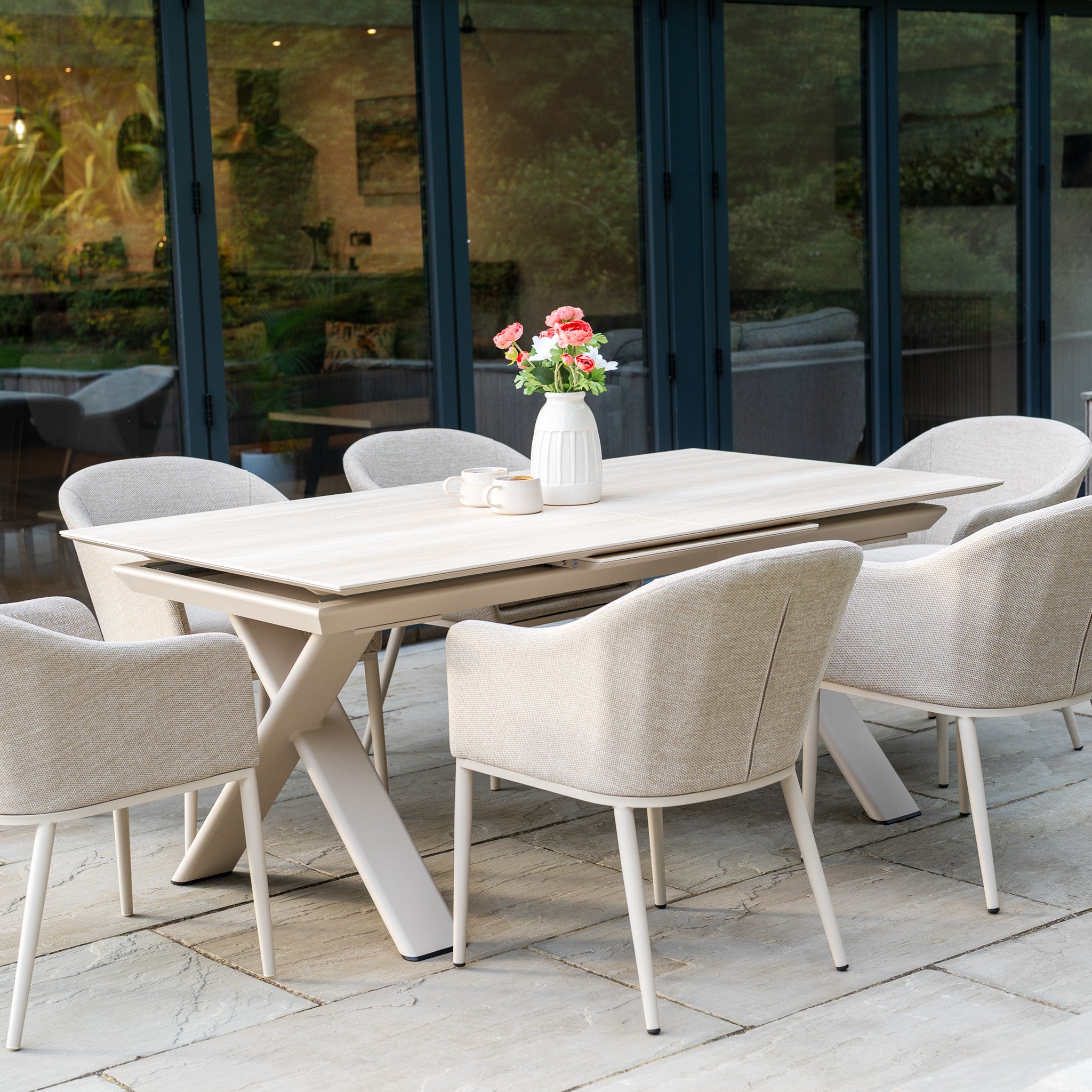 The Luna 6 Seat Outdoor Fabric Dining Set with Albatross Extending Table in Fawn includes six beige chairs with all-weather fabric and a light wood extending table, completed with a vase of flowers on top.
