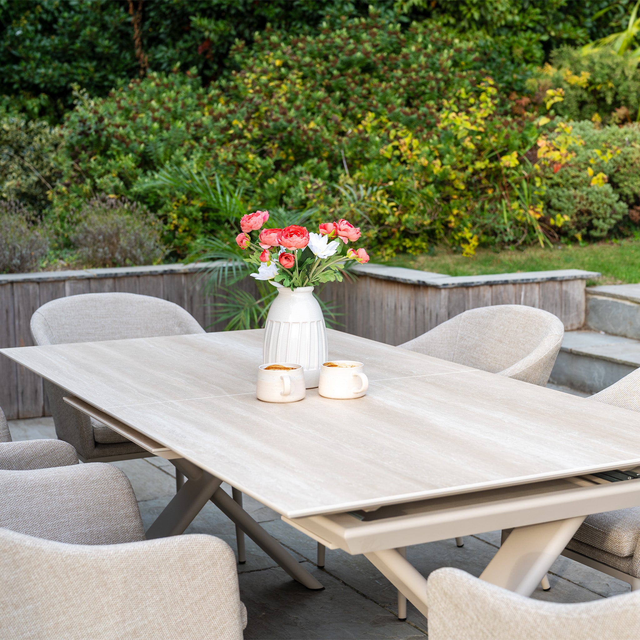 The Luna 6 Seat Outdoor Fabric Dining Set with Albatross Extending Table in Fawn, styled with pink flowers and mugs, is set in a garden.