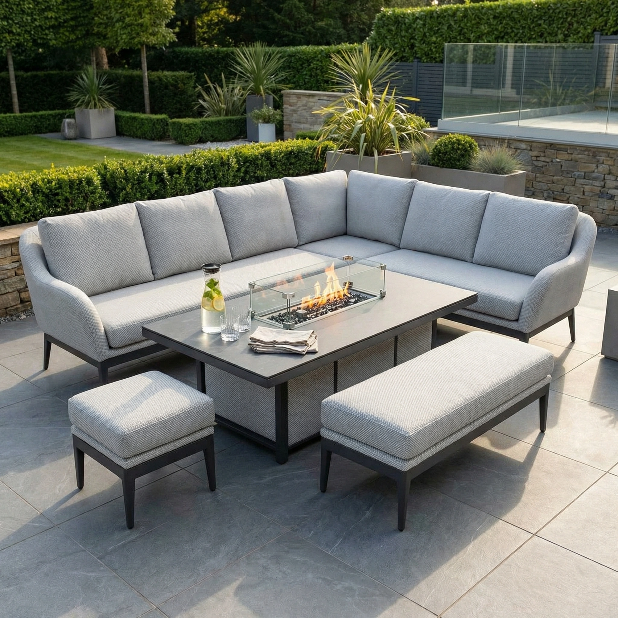 The Luna Outdoor Fabric Rectangular Corner Dining Set with Rising Firepit Table in Oyster Grey (Right Hand) features stylish fabric cushions and a rising firepit table, perfect for outdoor dining among lush plants and greenery.
