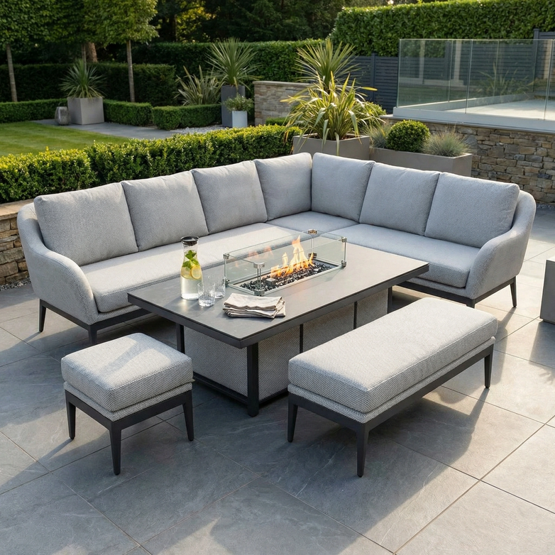 The Luna Outdoor Fabric Rectangular Corner Dining Set with Rising Firepit Table in Oyster Grey (Right Hand) features stylish fabric cushions and a rising firepit table, perfect for outdoor dining among lush plants and greenery.