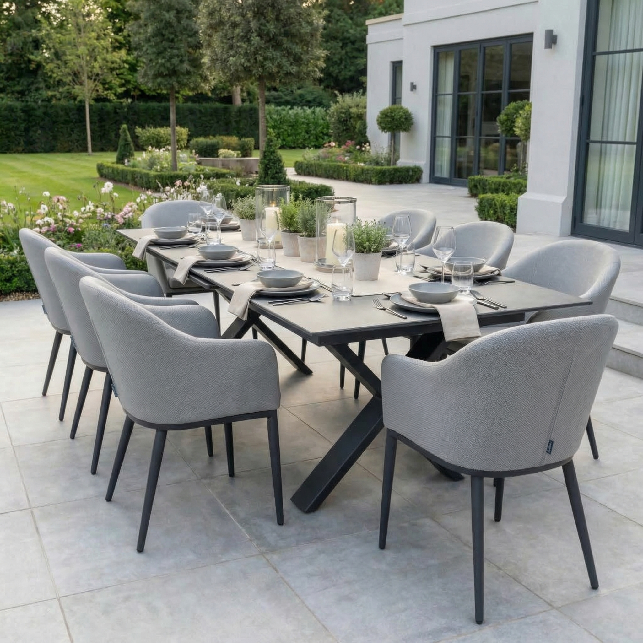 The Luna 8 Seat Outdoor Fabric Dining Set with Albatross Extending Table in Oyster Grey is styled for dining on a patio beside a garden.