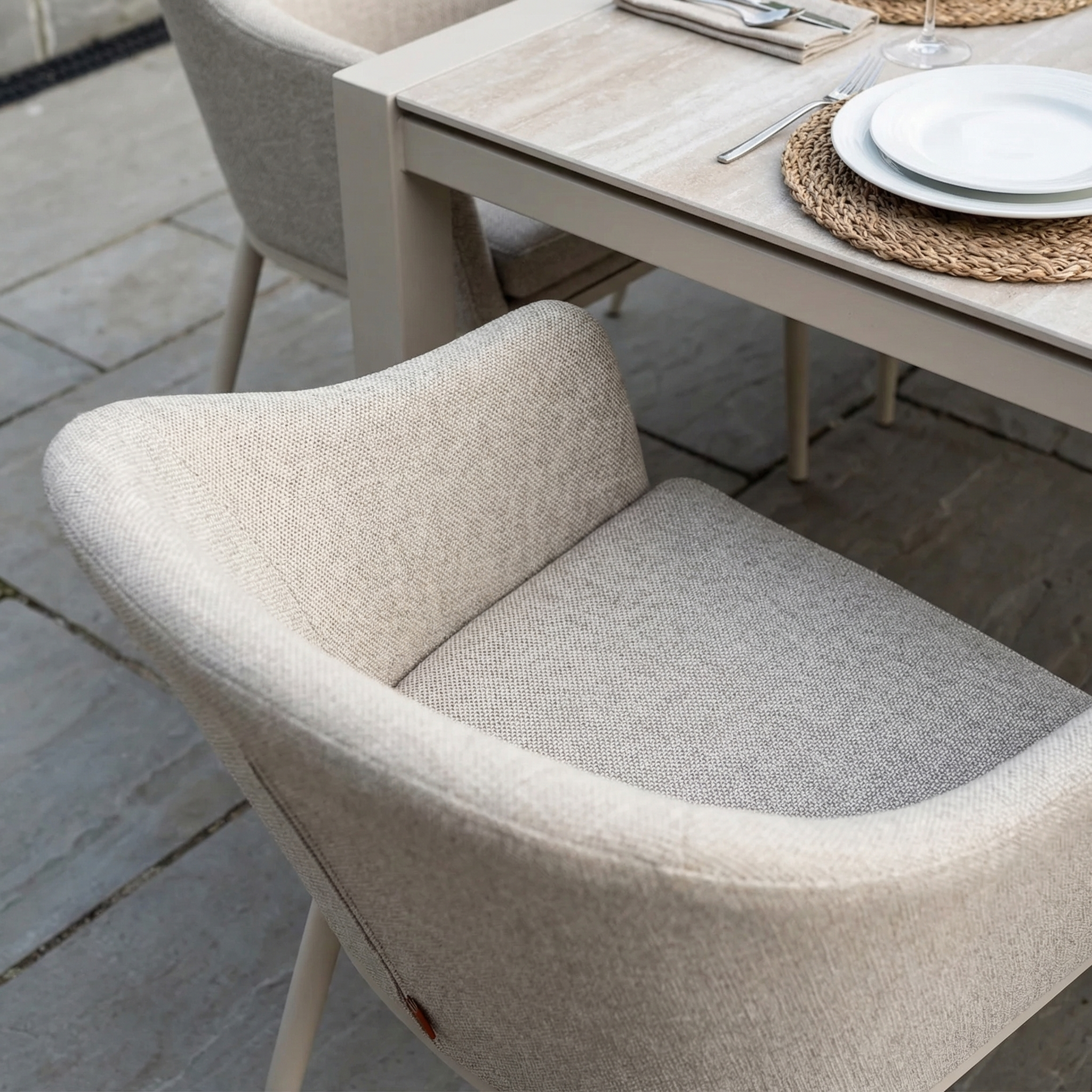 The Luna 8 Seat Outdoor Fabric Extending Ceramic Dining Set in Fawn is arranged with plates, cutlery, and woven placemats, complemented by a beige all-weather upholstered chair.