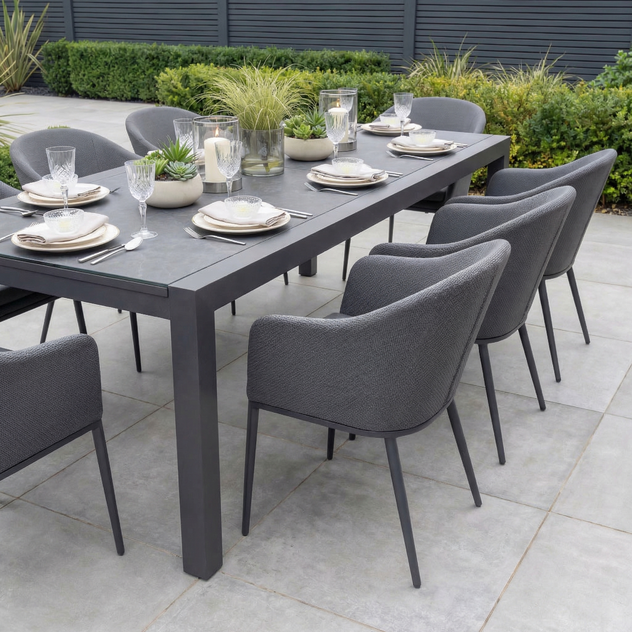 The Luna 8 Seat Outdoor Fabric Extending Ceramic Dining Set in Grey features eight gray chairs, a stain-resistant table, and is set with plates, glasses, and potted plants.